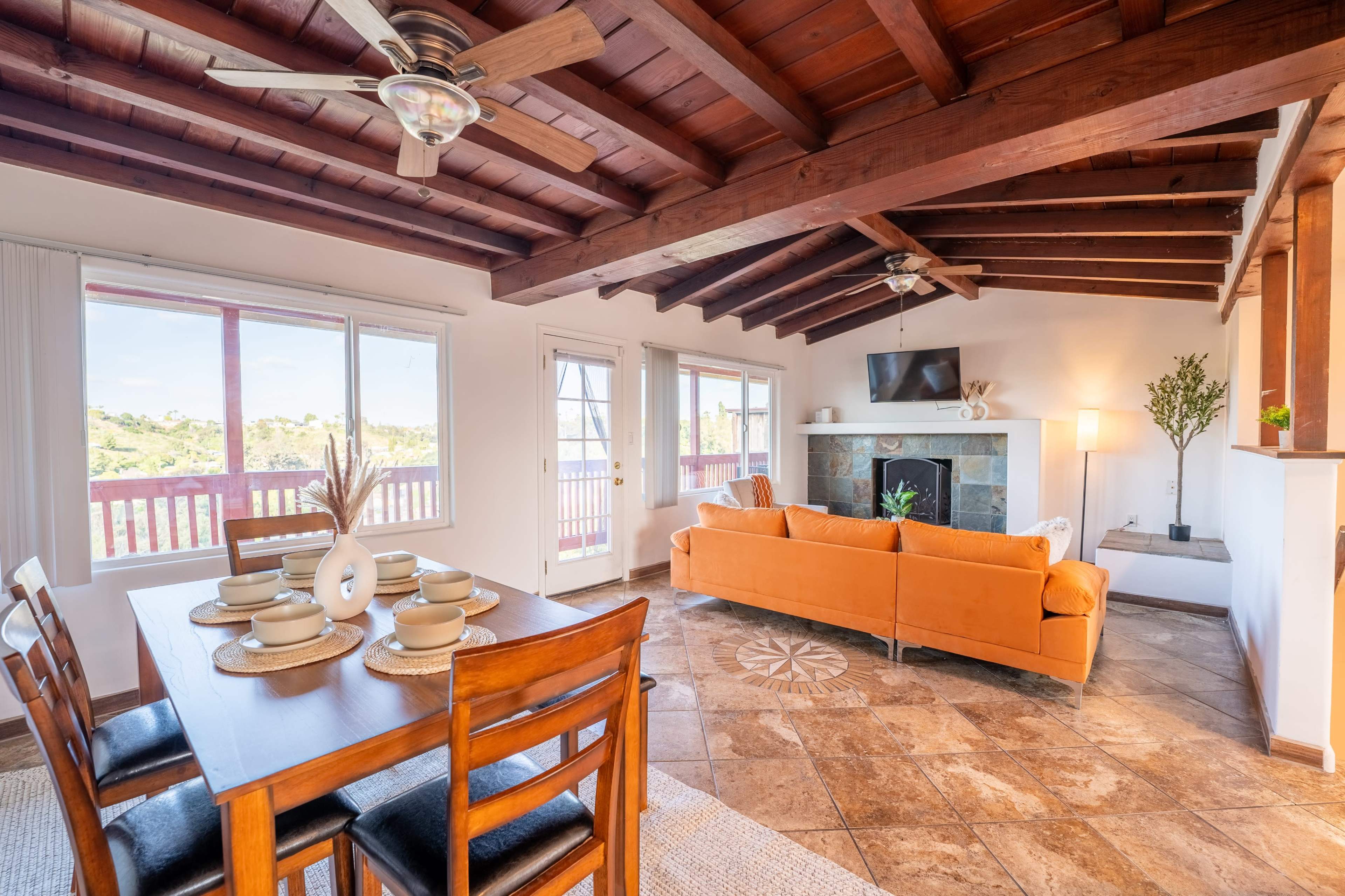 The image shows a living and dining area with a wooden ceiling, a table set for dining, an orange sofa, and large windows offering views of the outdoors.