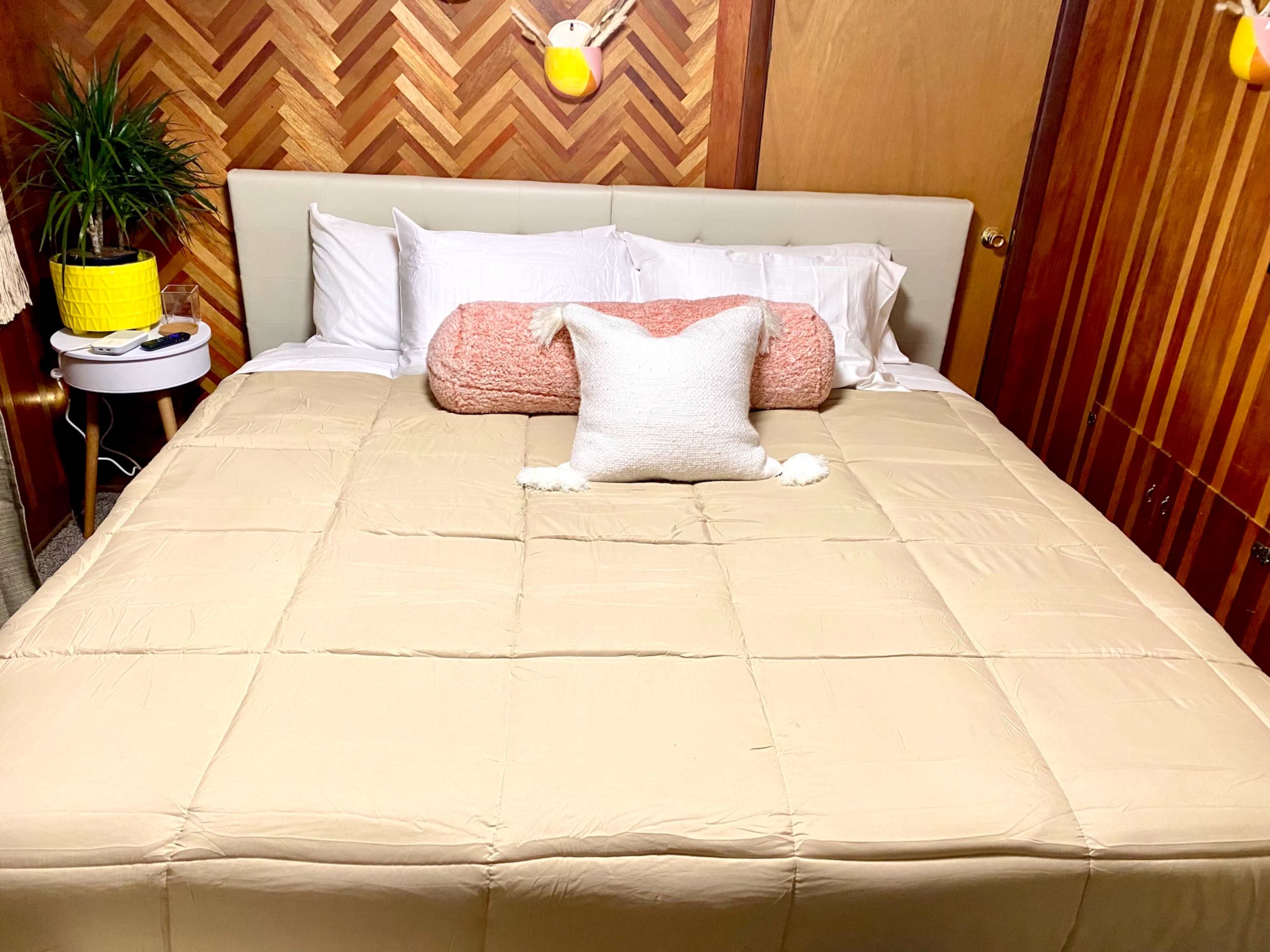 A neatly made bed with a beige comforter, decorative pillows, and a nightstand beside it in a room with wooden walls.