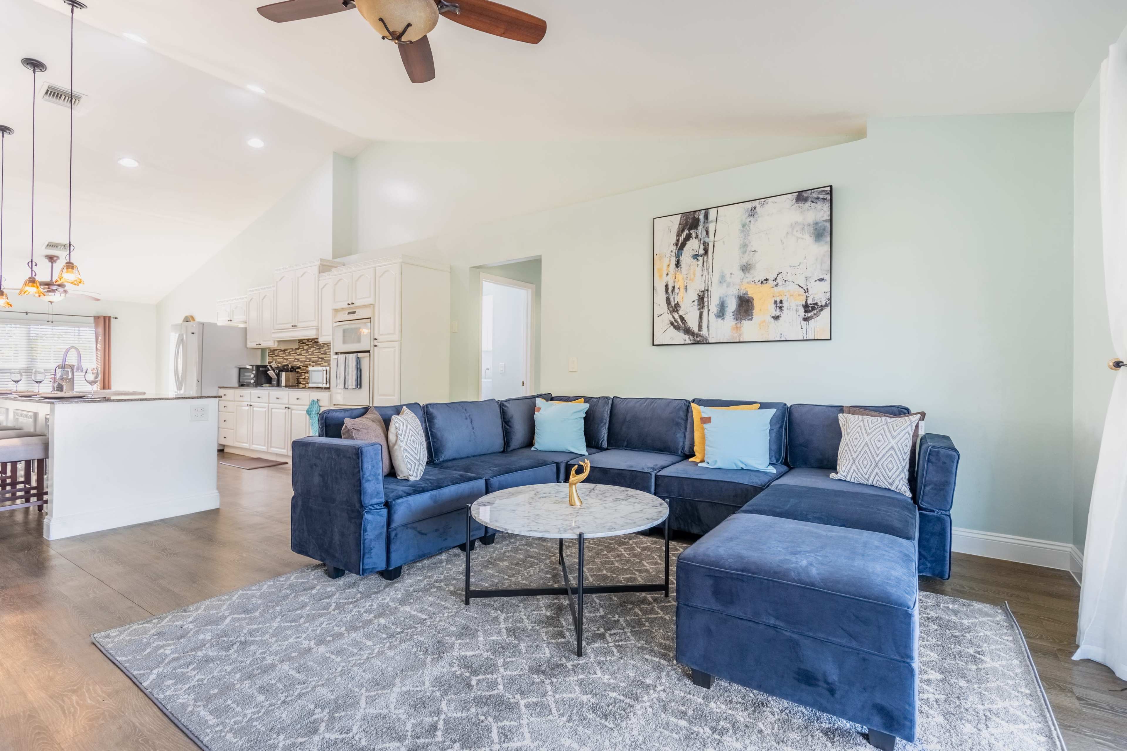 A blue sectional sofa with colorful cushions is arranged around a circular coffee table on a patterned rug in a light-filled living area that opens to a kitchen.