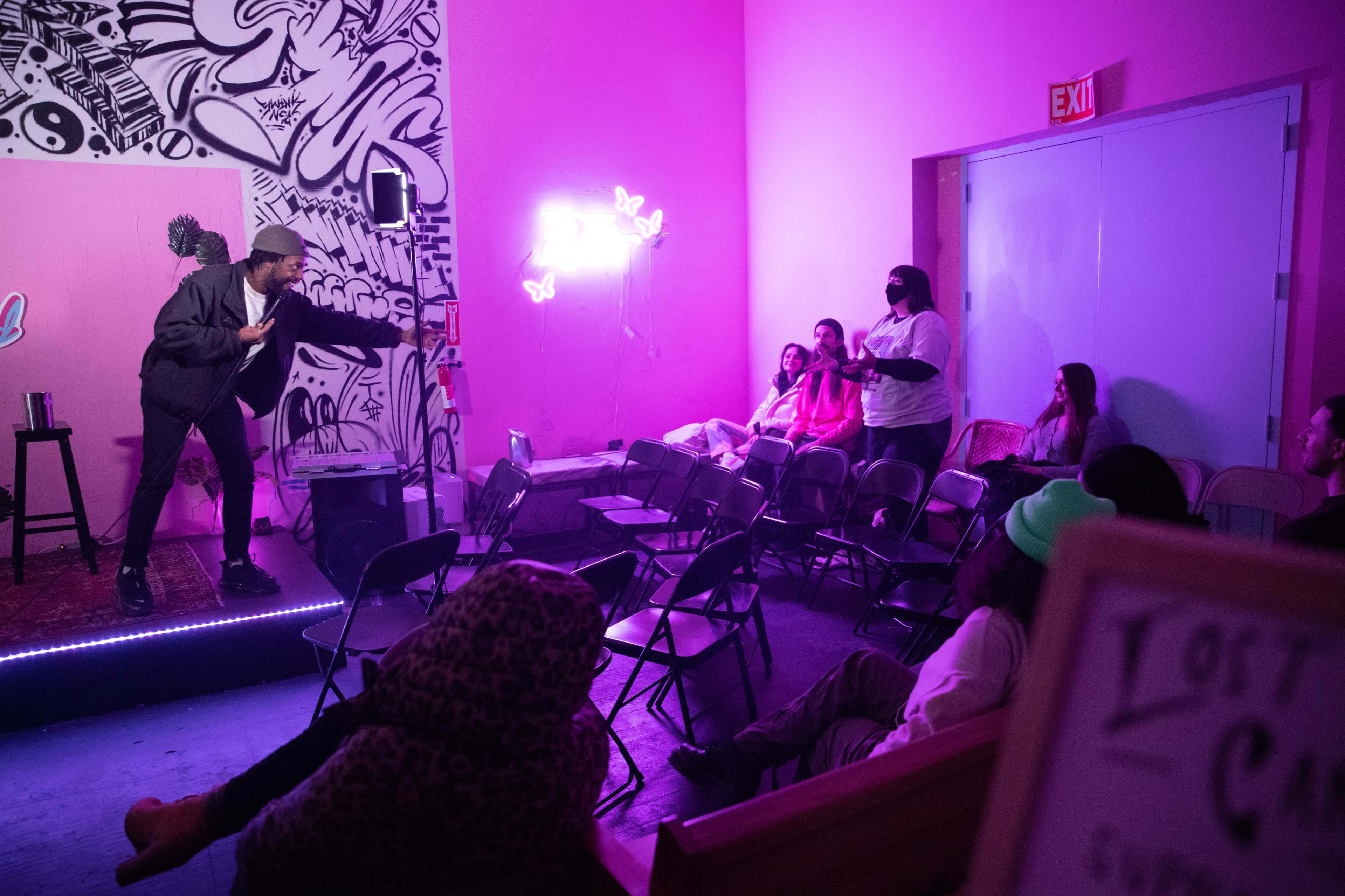 A performer stands on a small stage in a brightly lit room, while an audience sits in folding chairs, some facing the stage and others engaged in conversation.