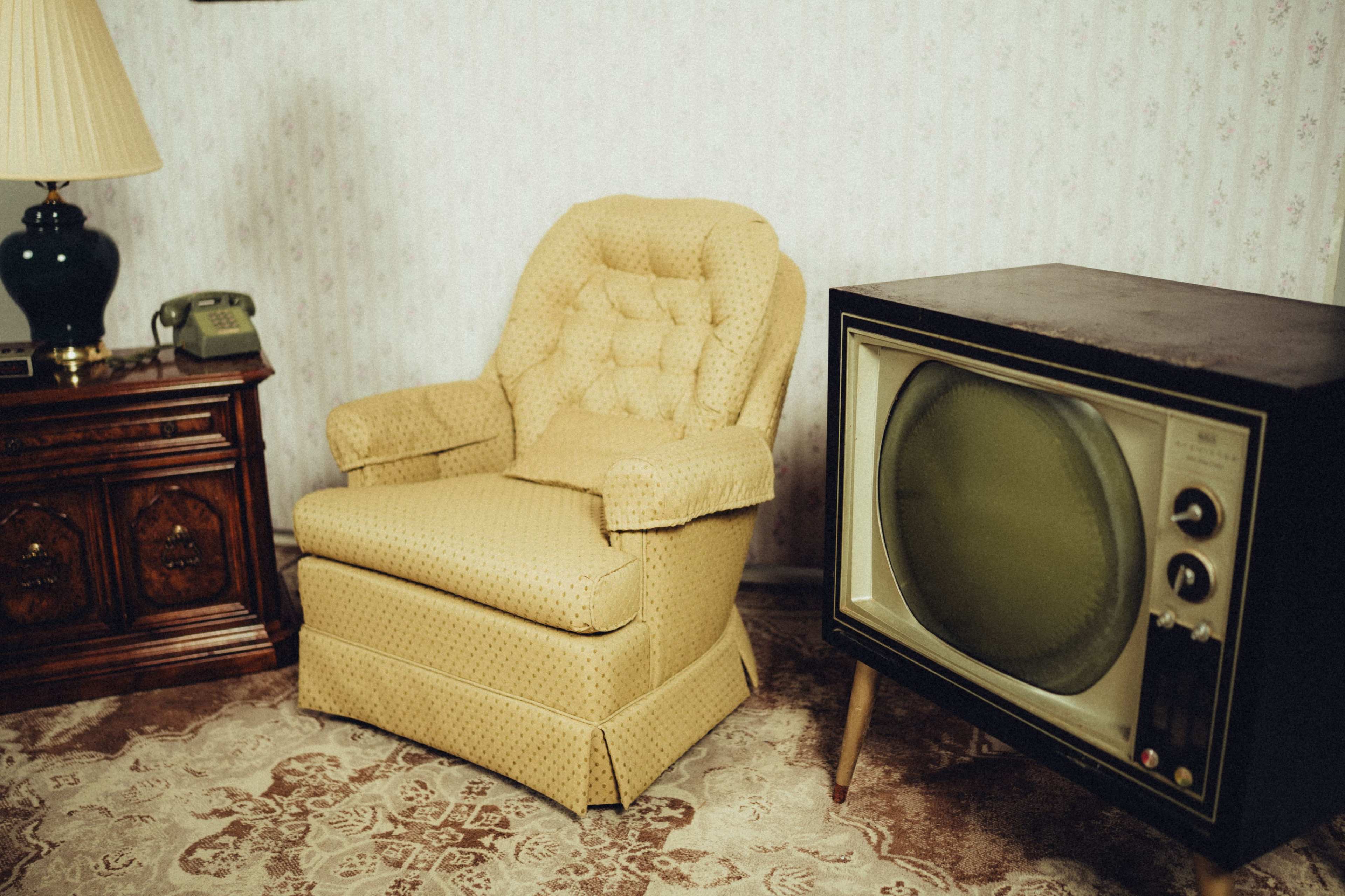 The scene features a vintage living room with a patterned yellow armchair, an old television set, a wooden side table with a lamp, and a telephone.