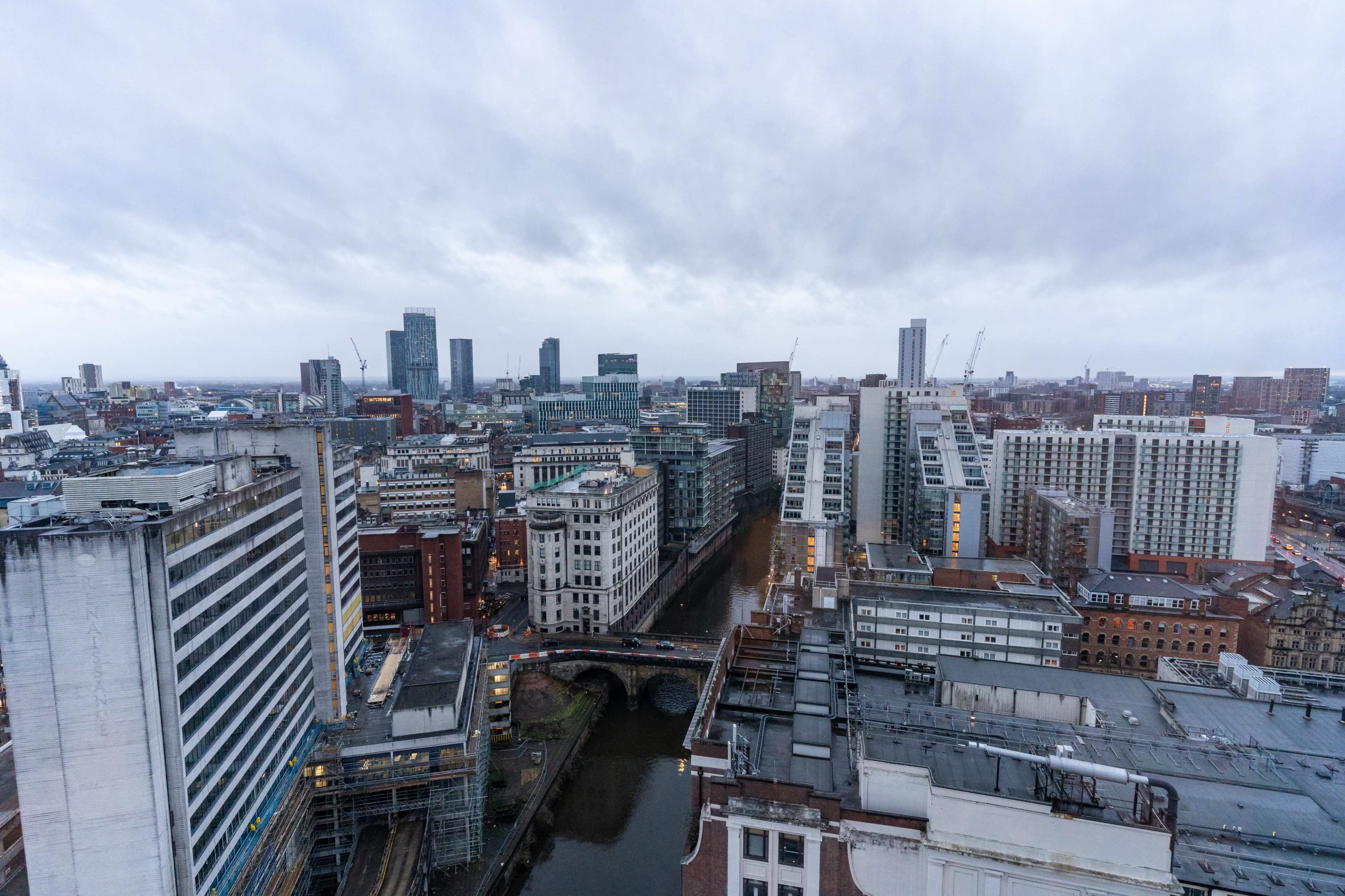 An aerial view of a cityscape with a river running through, surrounded by a mix of modern and older buildings under a cloudy sky.