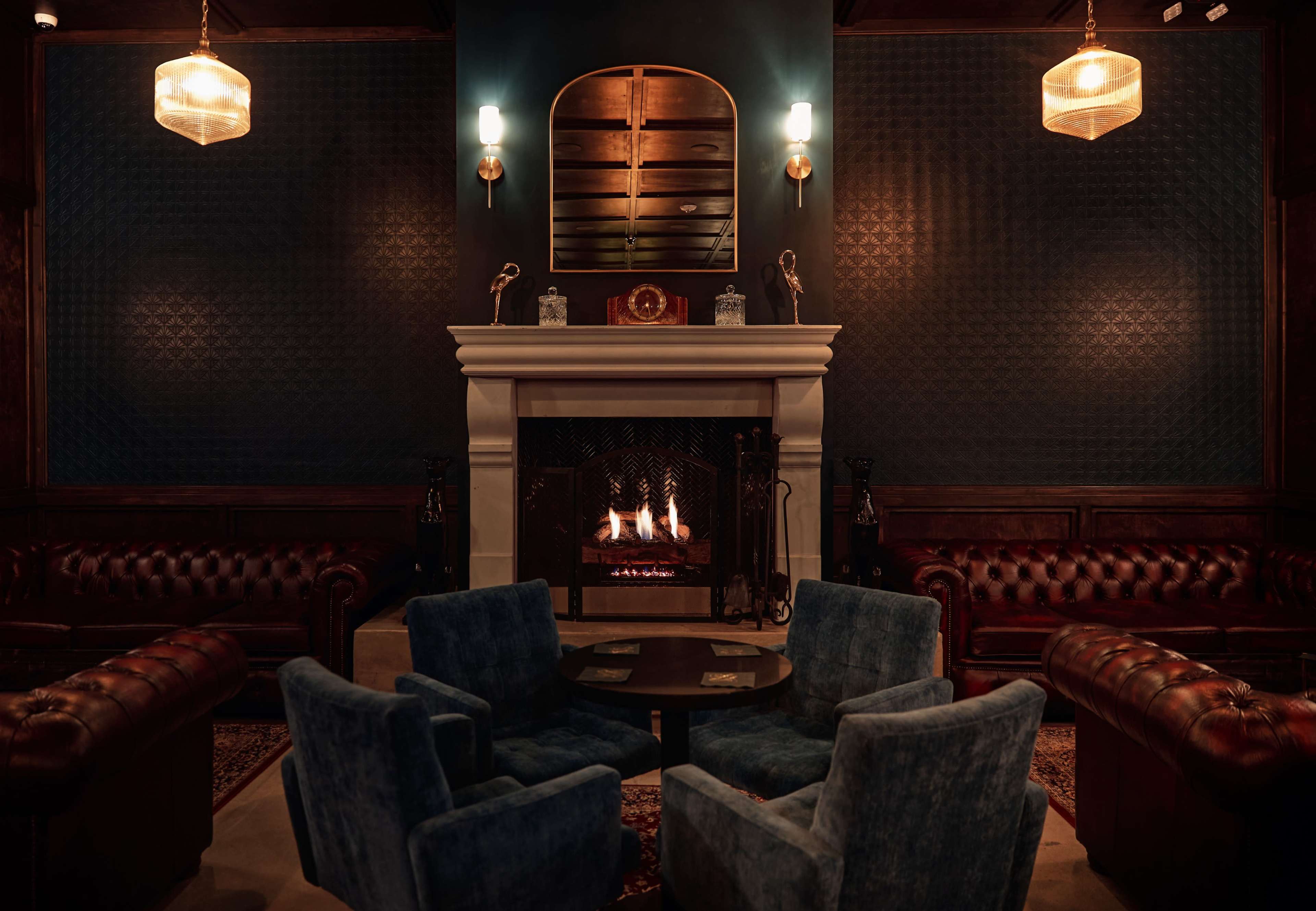 A cozy lounge features a fireplace surrounded by leather sofas and blue velvet chairs under warm lighting.