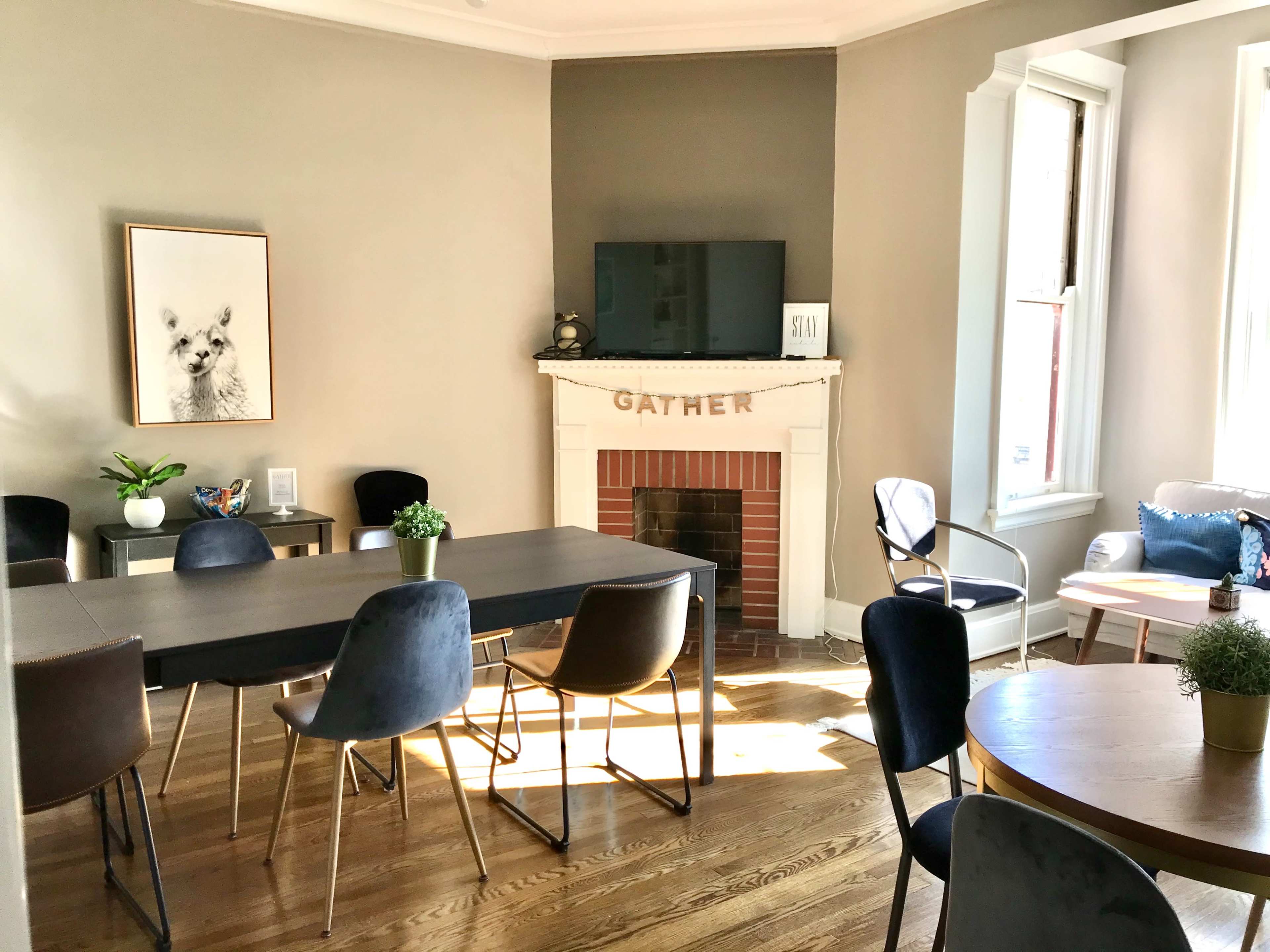 The image shows a well-lit room with a dining table, several chairs, and a fireplace adorned with the word "GATHER," featuring a television above it and plants in various spots.