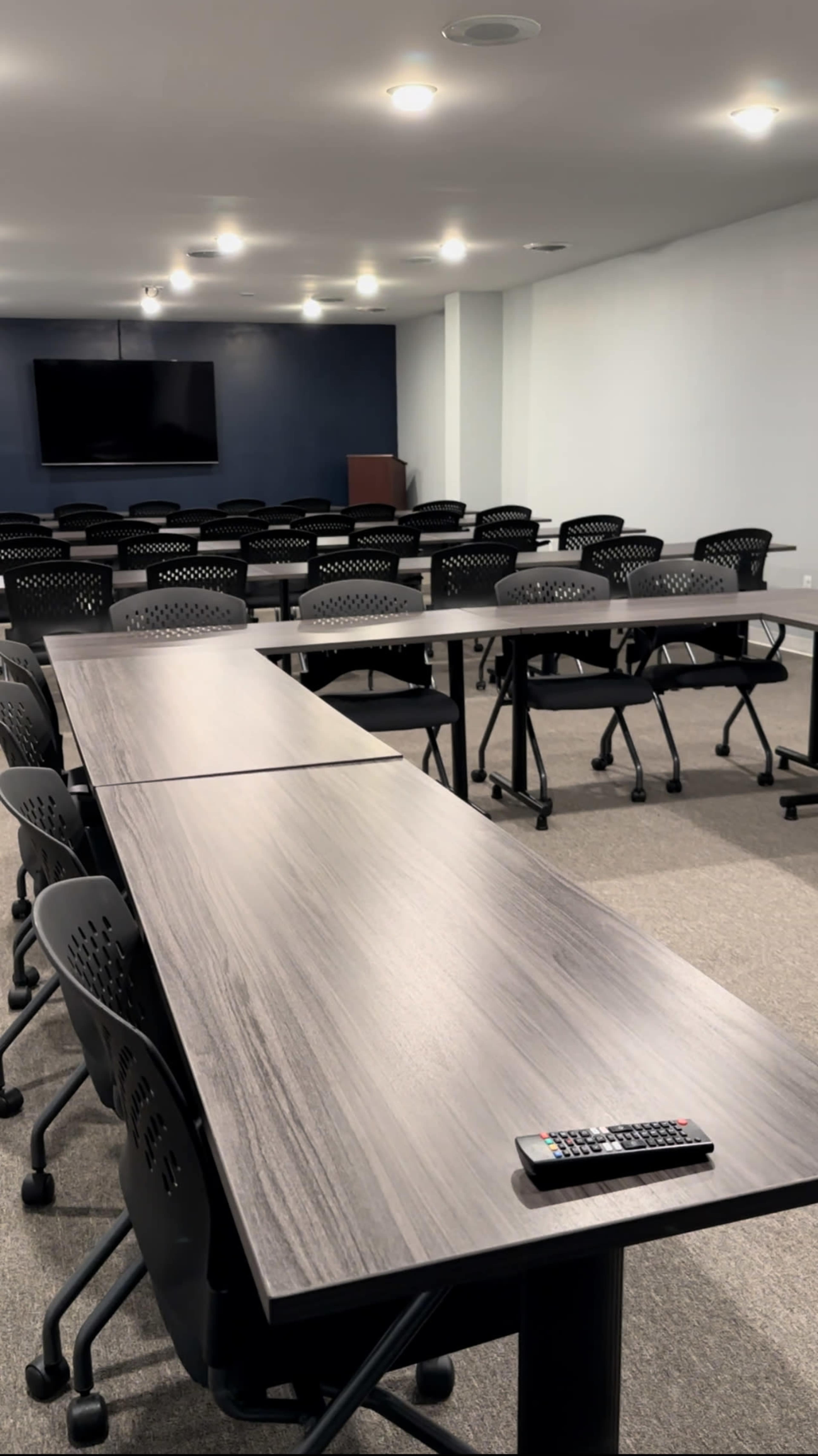 A conference room is set up with multiple rows of tables and chairs, facing a large screen on the wall.