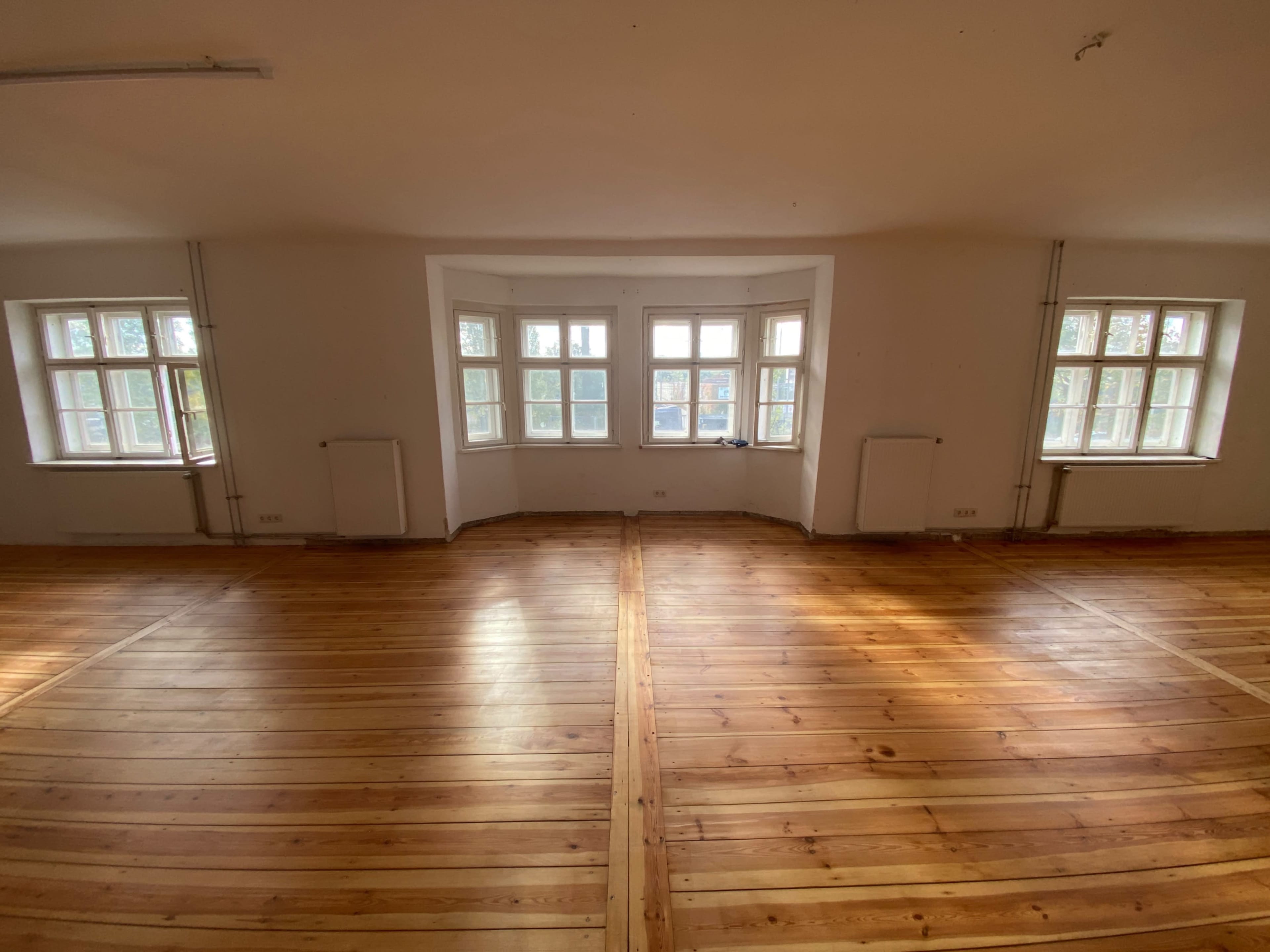 The image shows a spacious room with wooden floors and large windows arranged in a bay format along one wall.