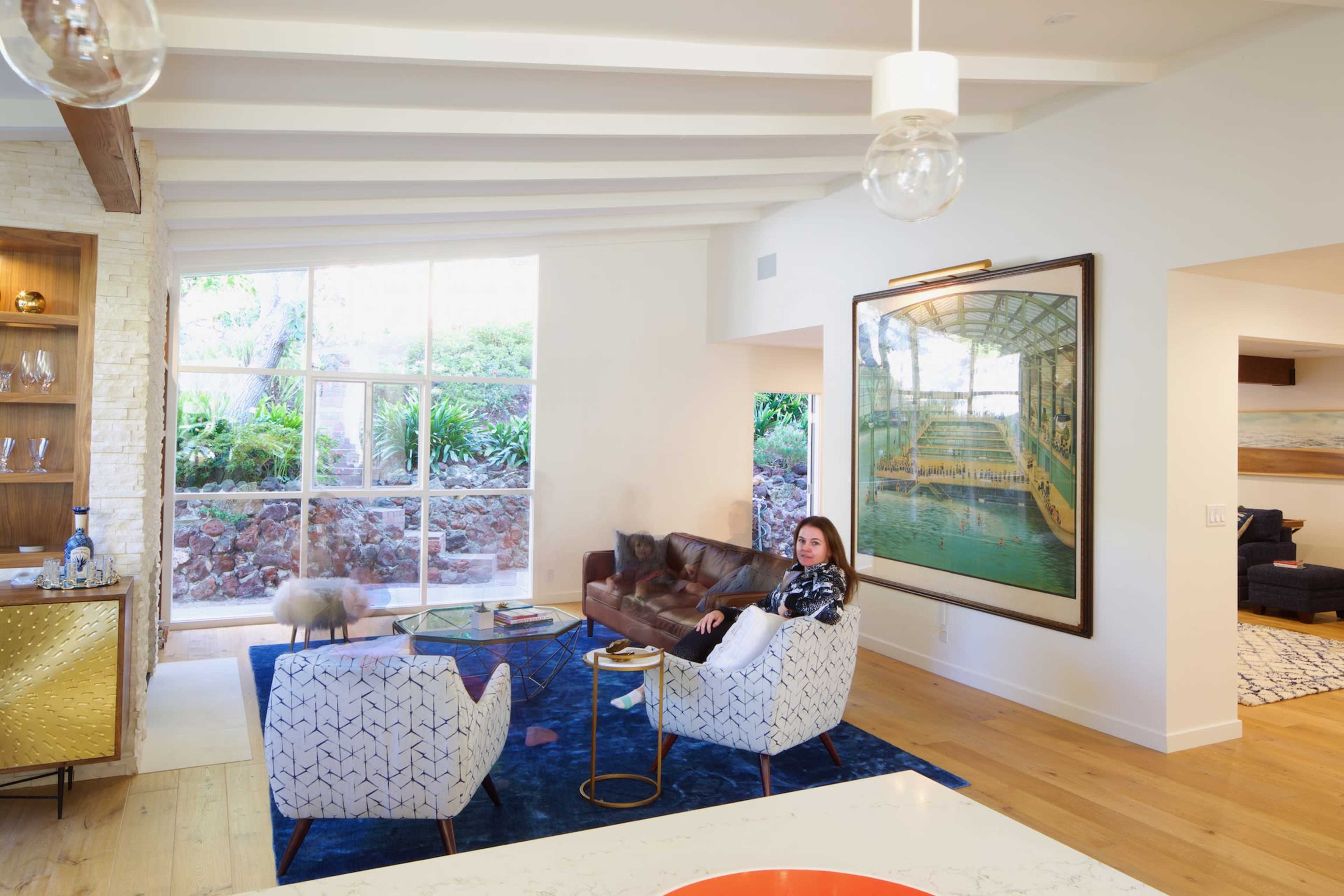 A woman sits in a modern living room with large windows and a colorful rug, surrounded by stylish furniture and artwork.