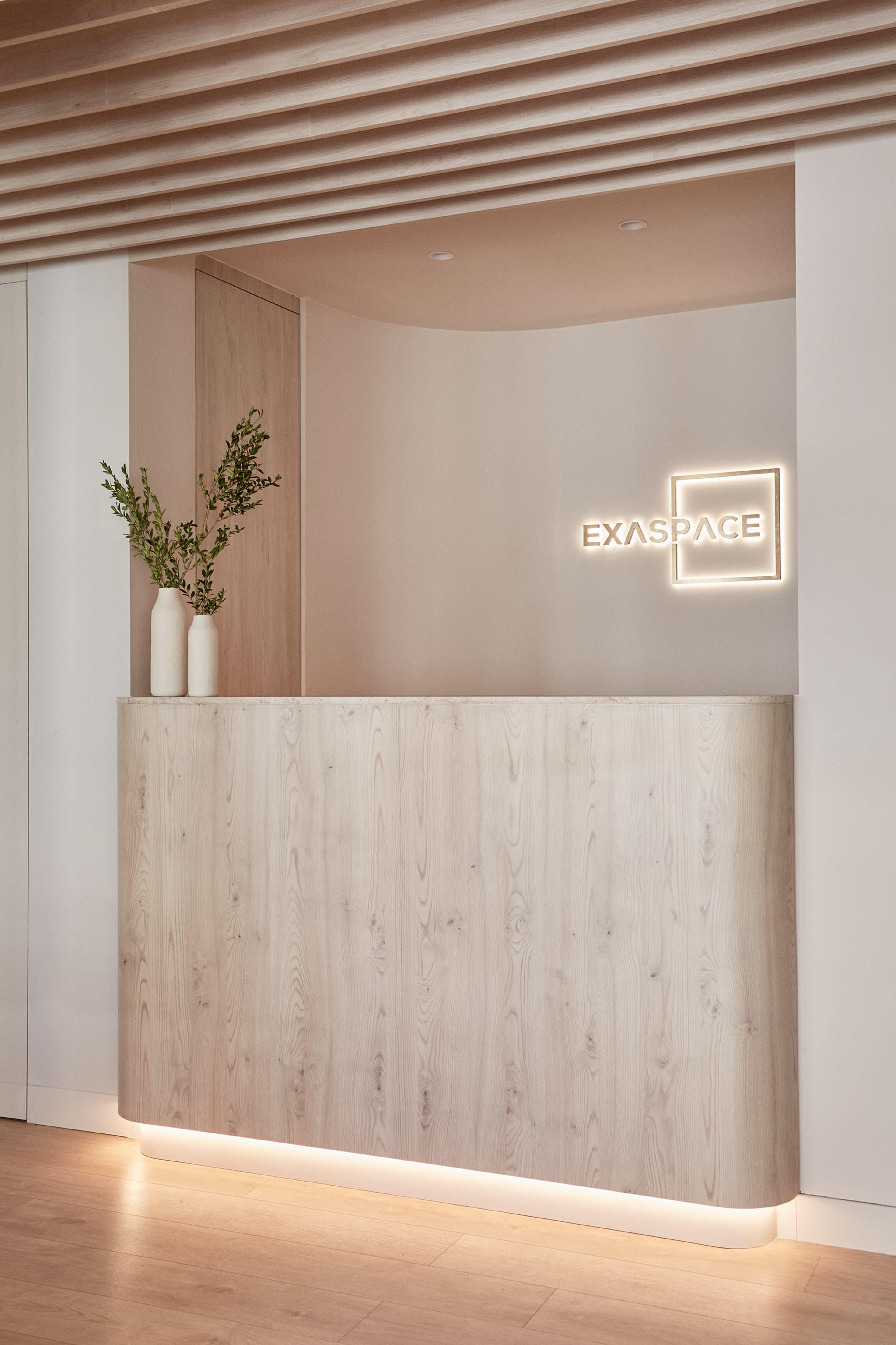 The image shows a modern reception desk with a light-colored wood finish, a minimalist vase with greenery, and a glowing sign that reads "EXASPACE."