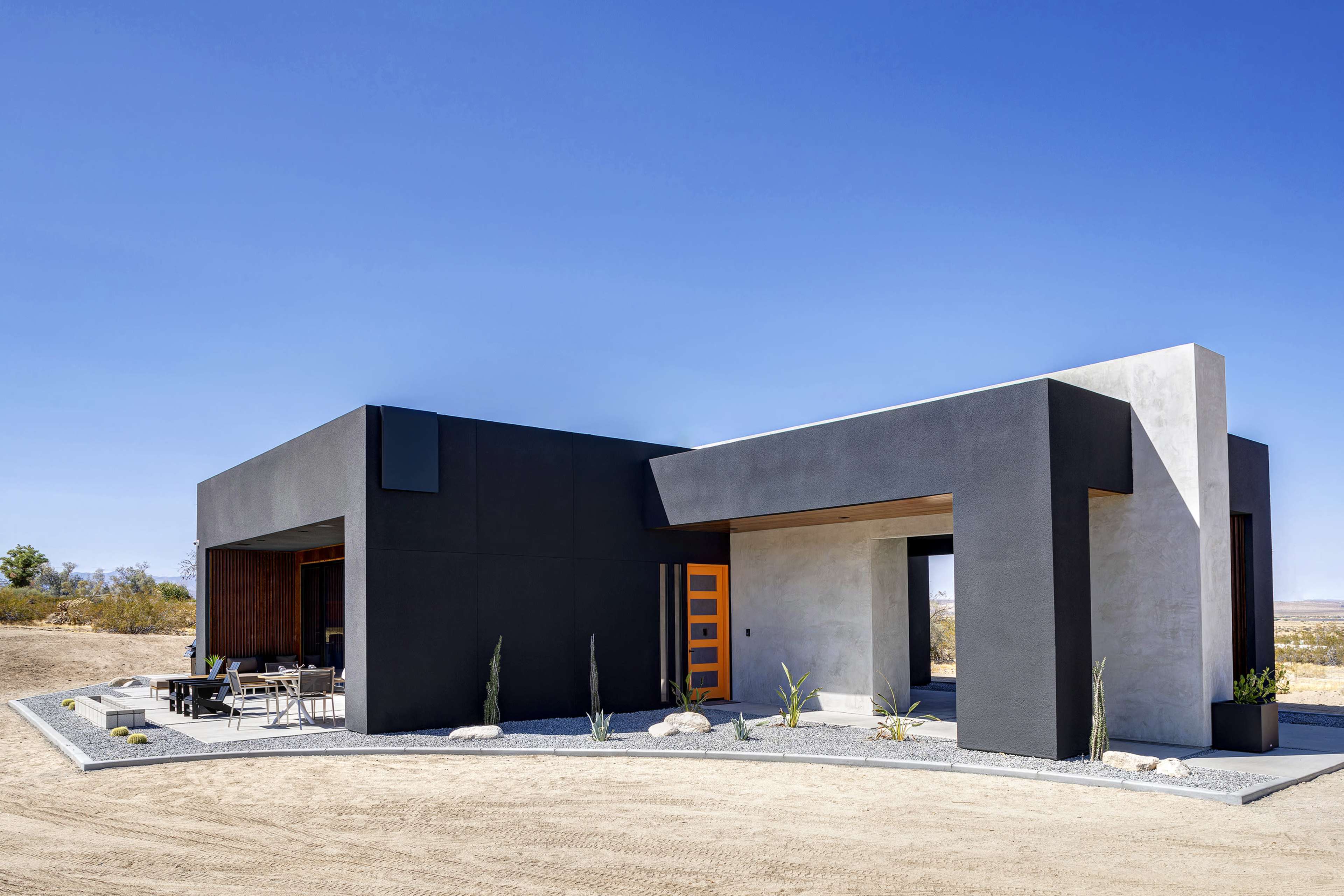A modern house with a contrasting black exterior and a bright orange door is set against a clear blue sky and a sparse landscape.
