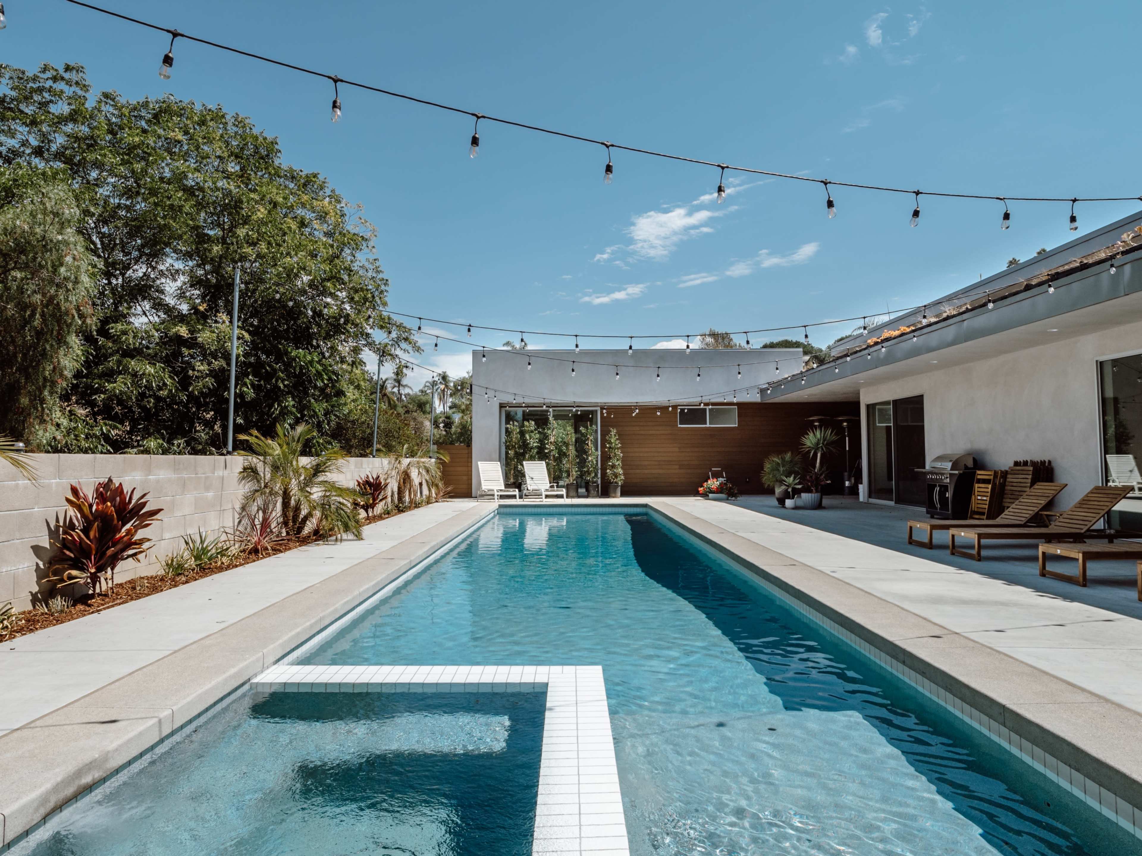 The image shows a modern backyard pool surrounded by lounge chairs and landscaped greenery, with string lights overhead.