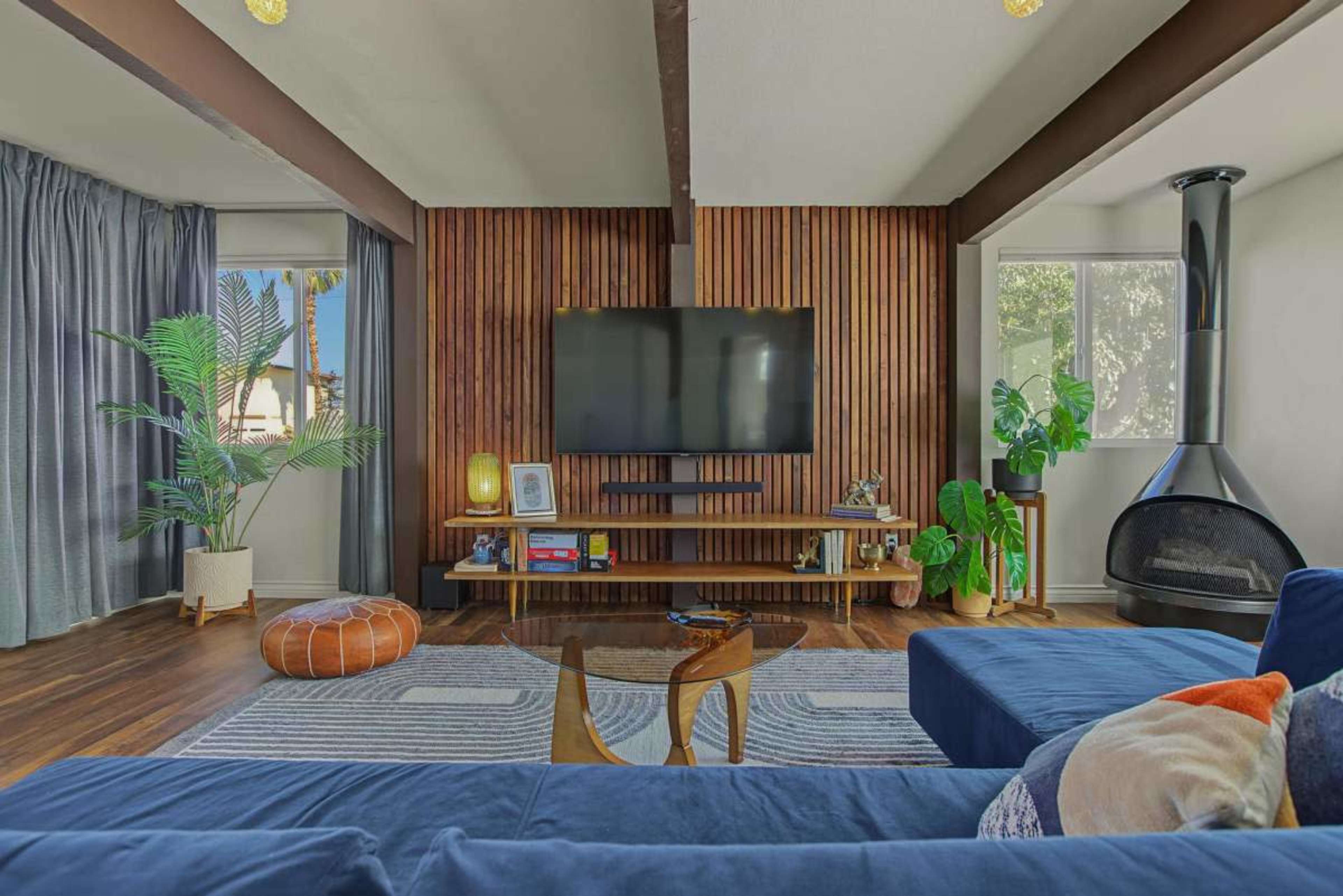 The image depicts a modern living room with a large television mounted on a wooden accent wall, a blue sofa, and a cozy seating area featuring a round pouf.