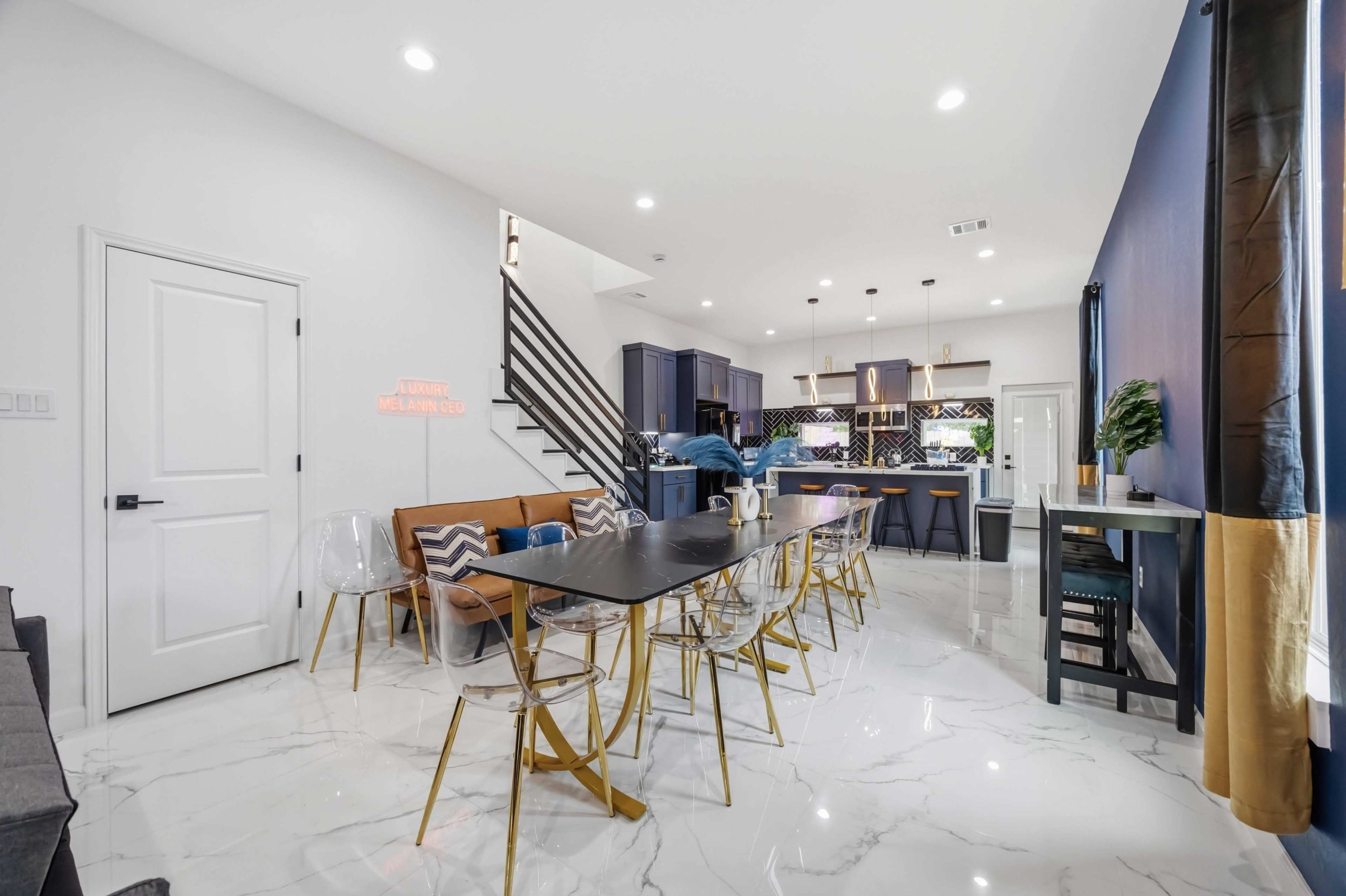 The image shows a modern dining area with a large table and chairs, adjacent to a kitchen featuring dark cabinetry and bright lighting.