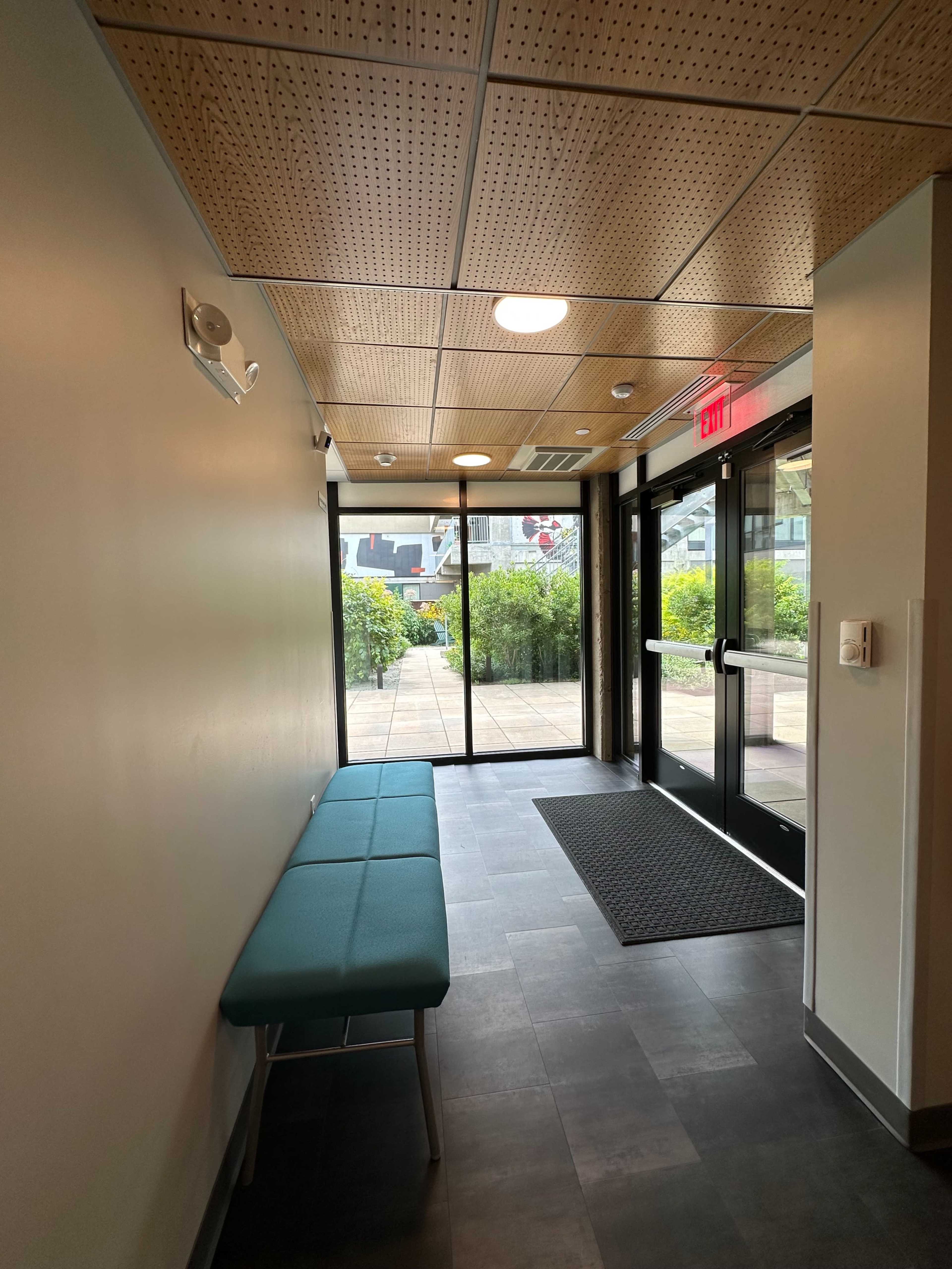 A hallway leading to large glass doors that open to an outdoor area, with a green bench along one wall.