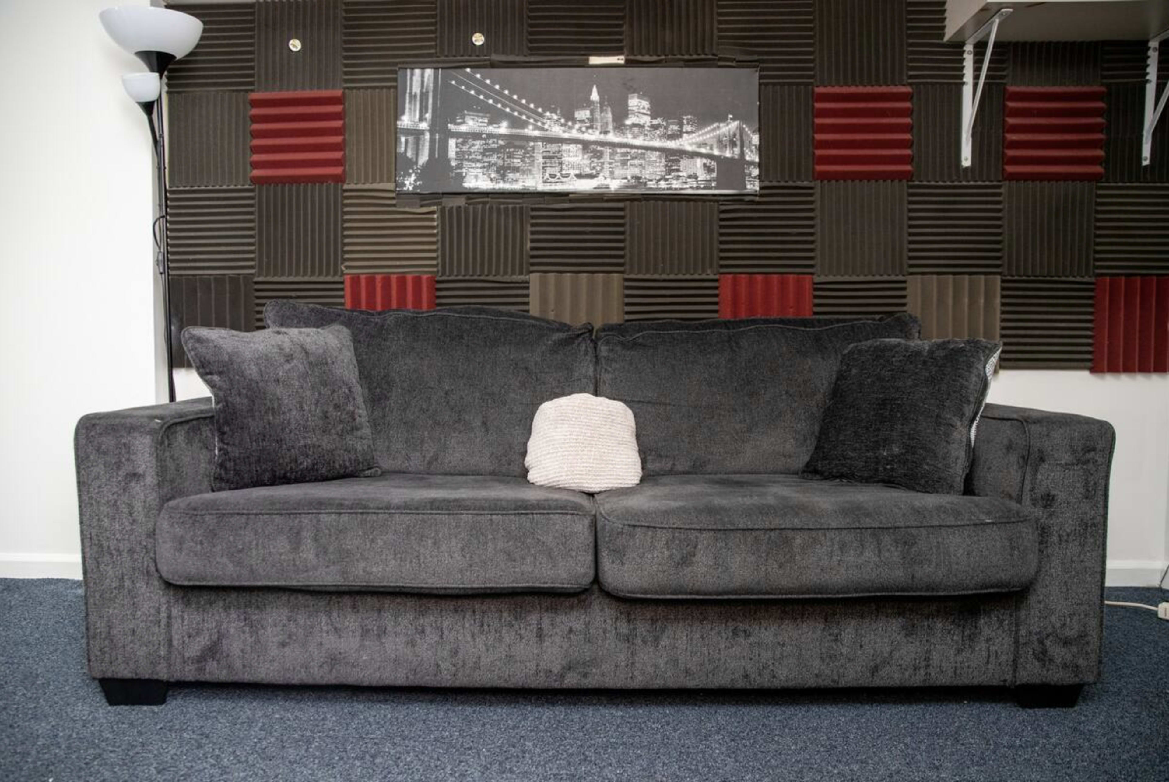 A gray sofa with two cushions and a small cream pillow is positioned against a wall featuring soundproof panels and a cityscape picture.