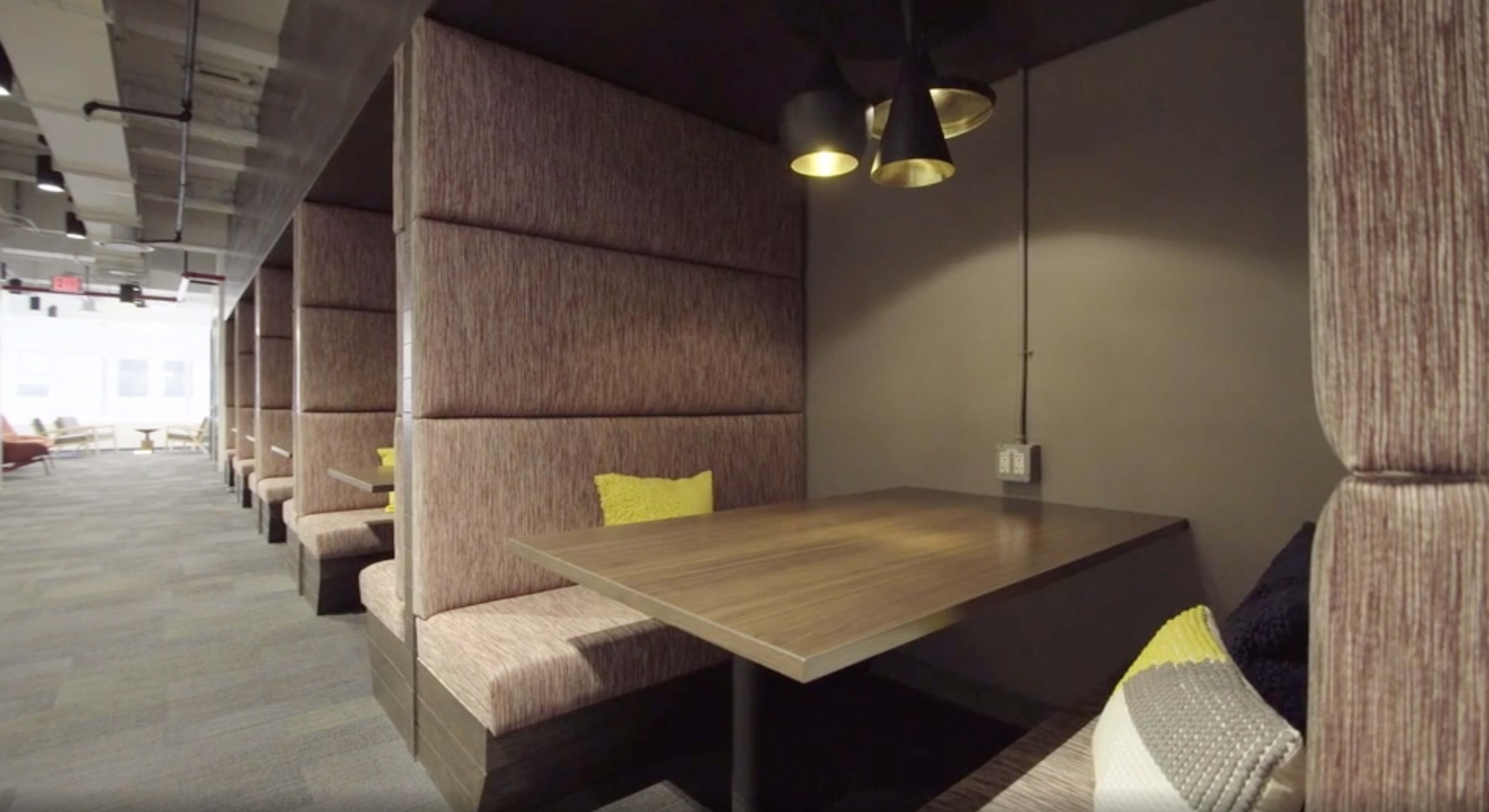 The image shows a modern office space with cozy seating areas featuring tall upholstered walls, wooden tables, and pendant lighting.