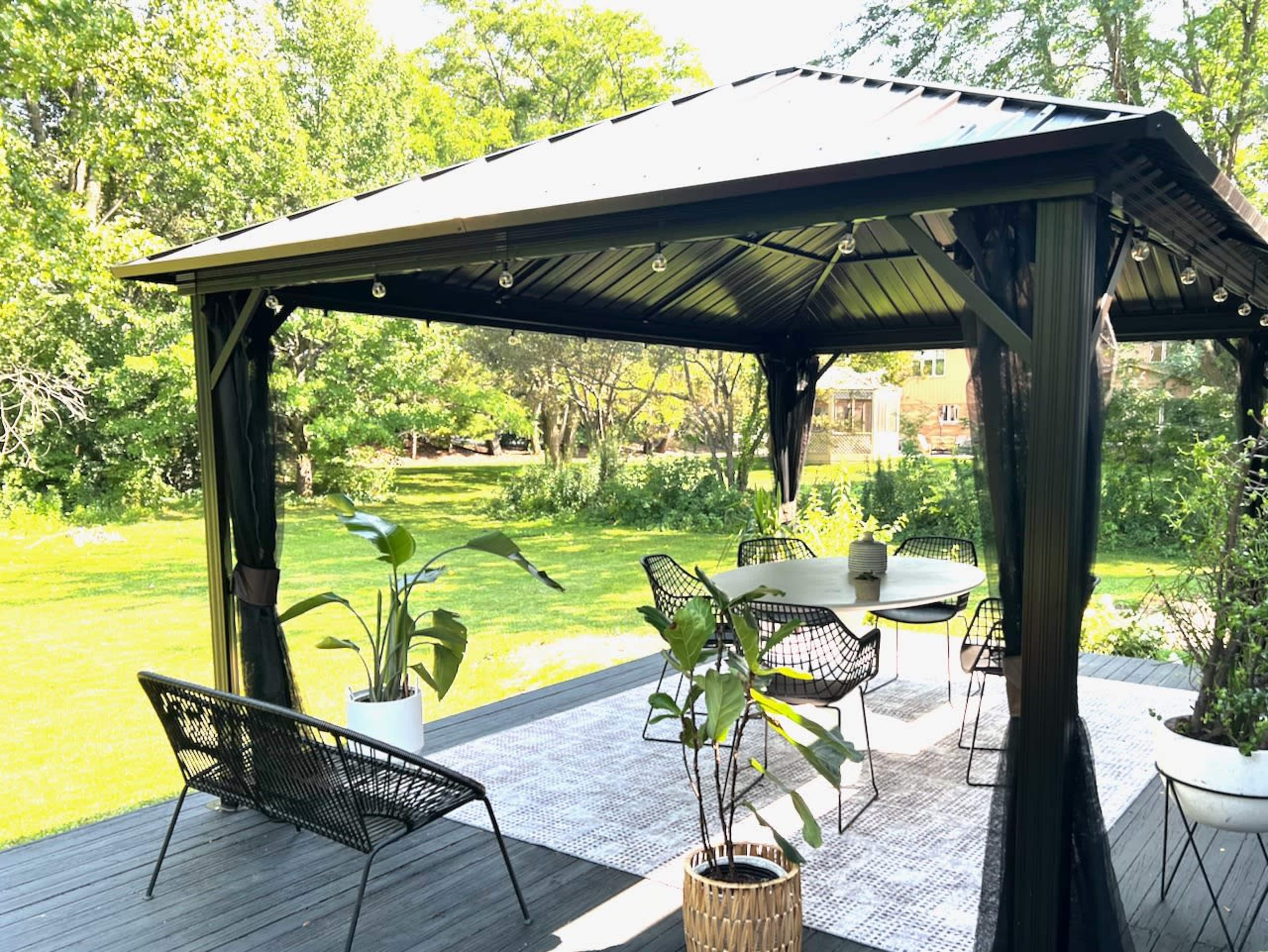 A gazebo with a dining table and chairs is set on a deck surrounded by greenery.