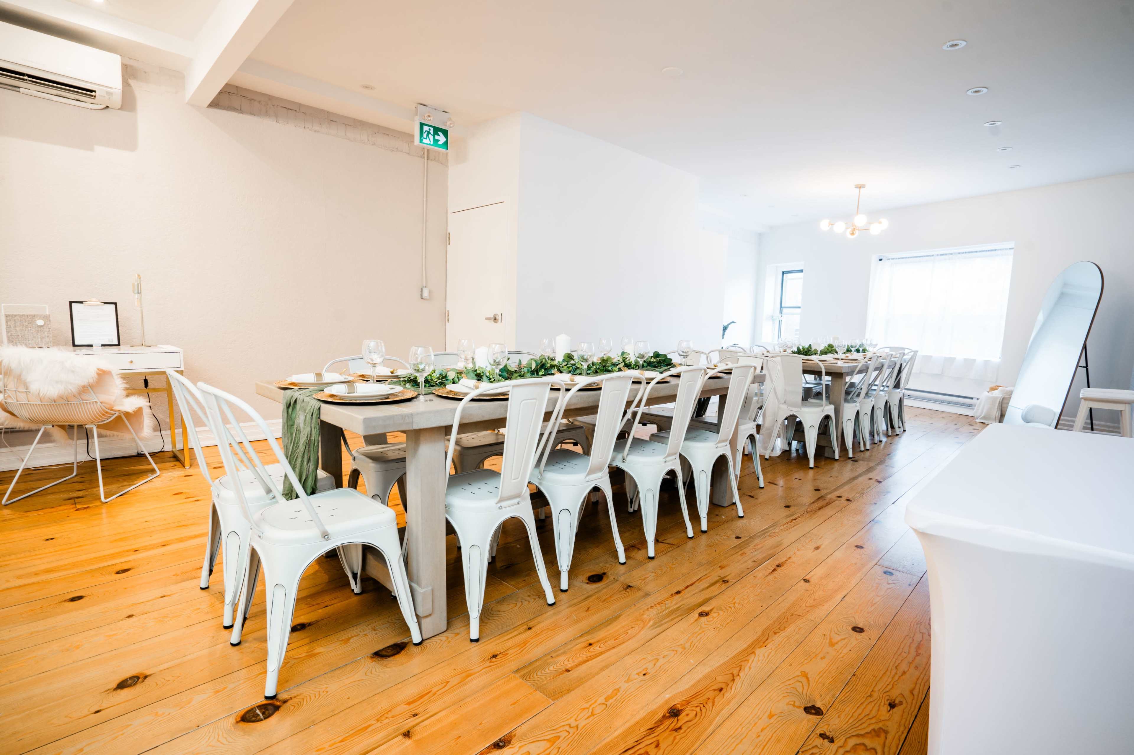 A long dining table is set with tableware and greenery in a spacious, well-lit room with wooden floors.
