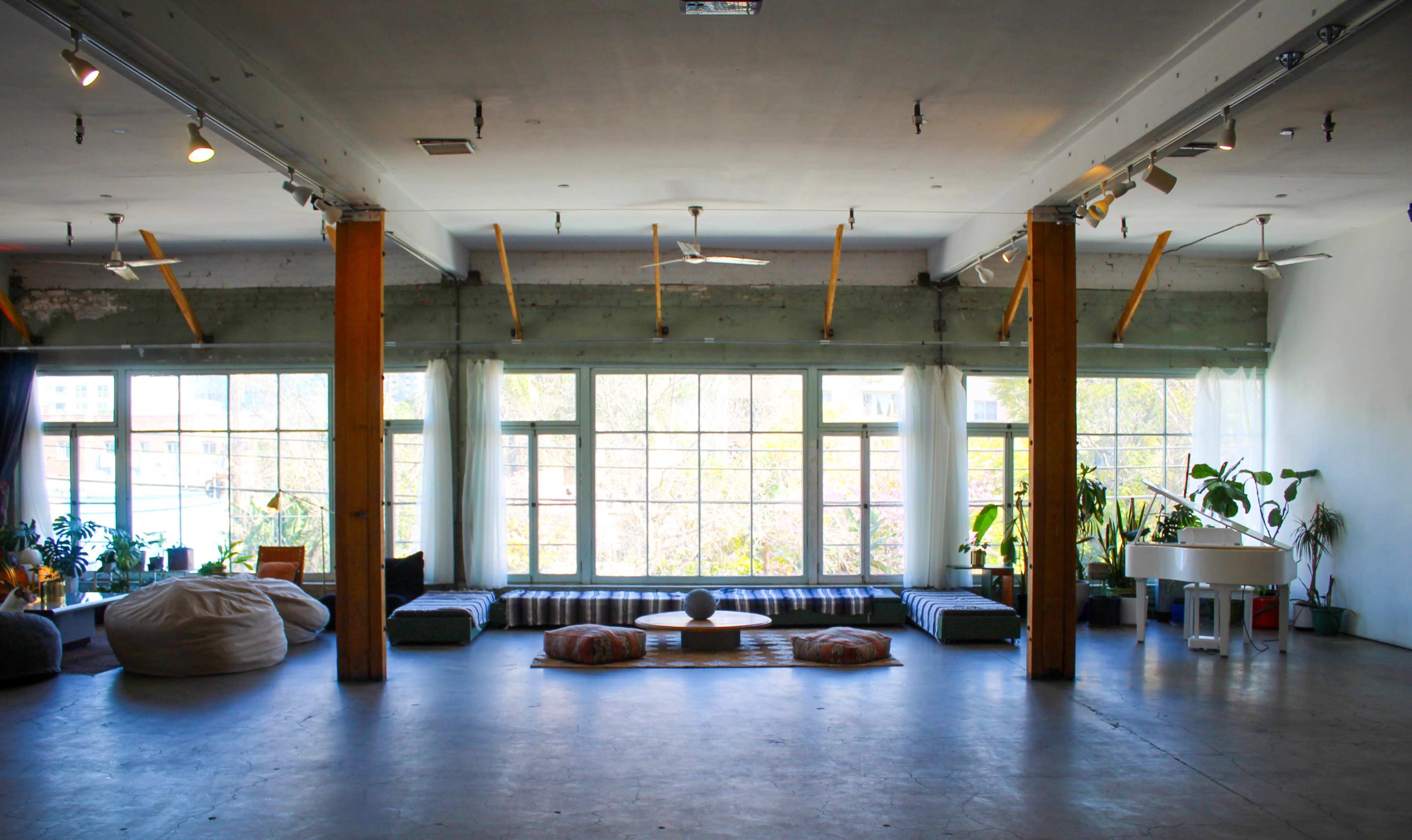The image shows a spacious interior with large windows, wooden beams, a minimalist seating area with cushions, and various indoor plants.