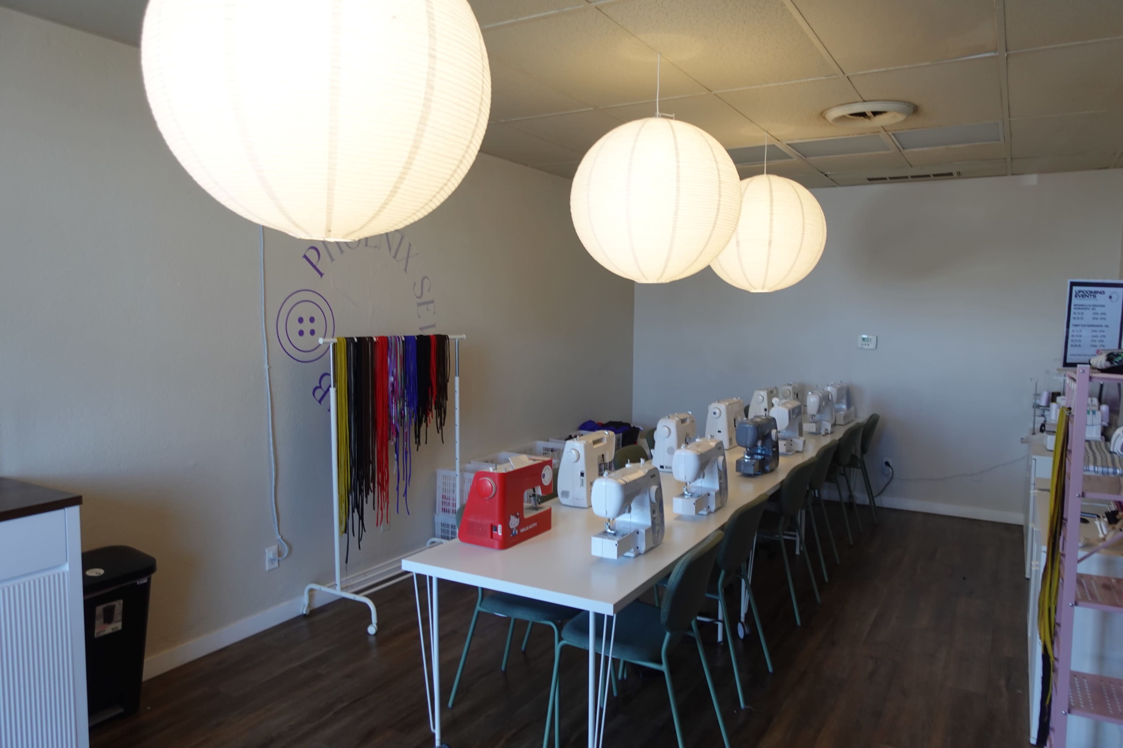 The image shows a bright sewing room with multiple sewing machines arranged on long tables under large round pendant lights.