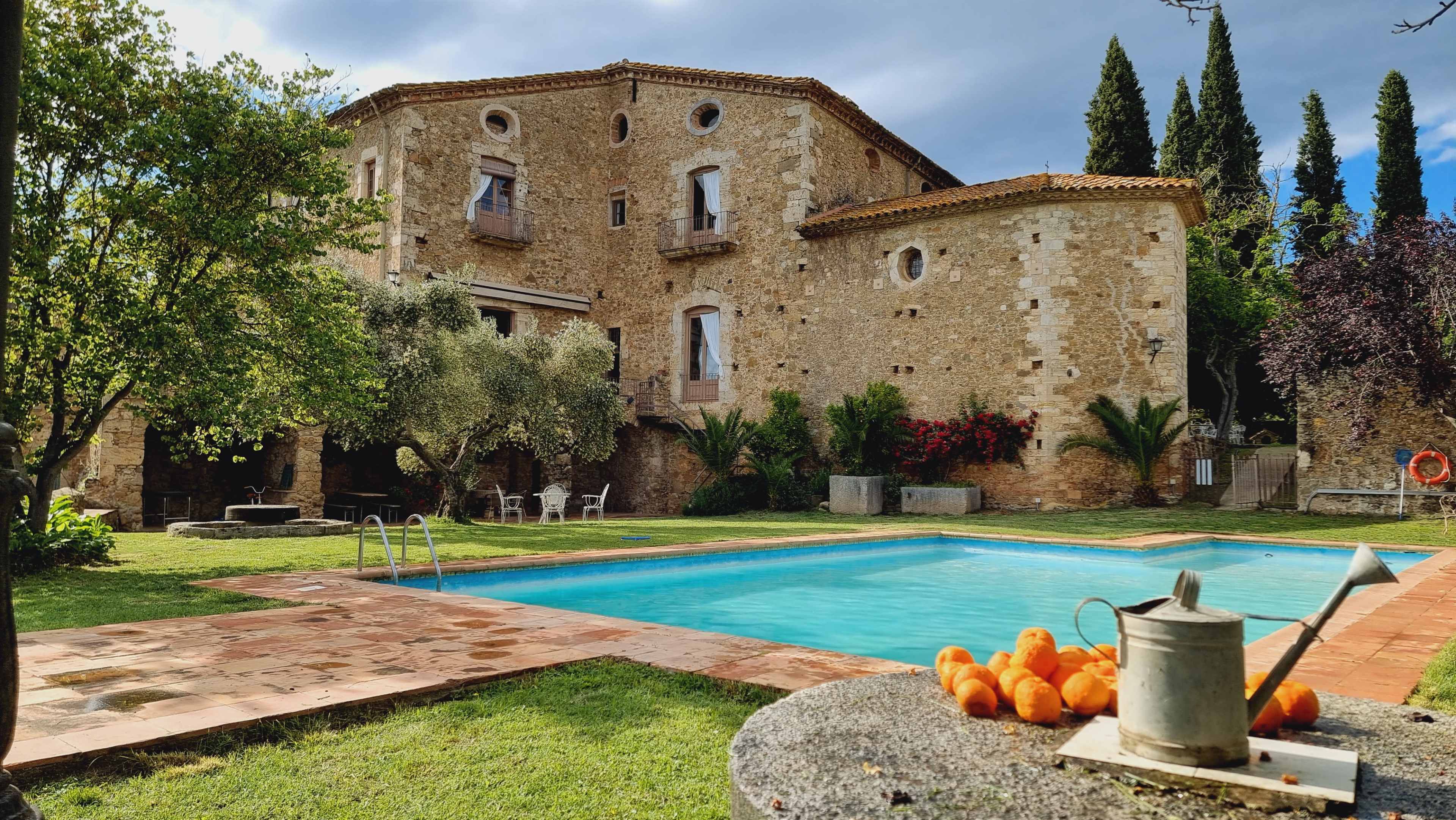 The image shows a stone villa surrounded by greenery and a swimming pool, with a stone well and a basket of oranges in the foreground.