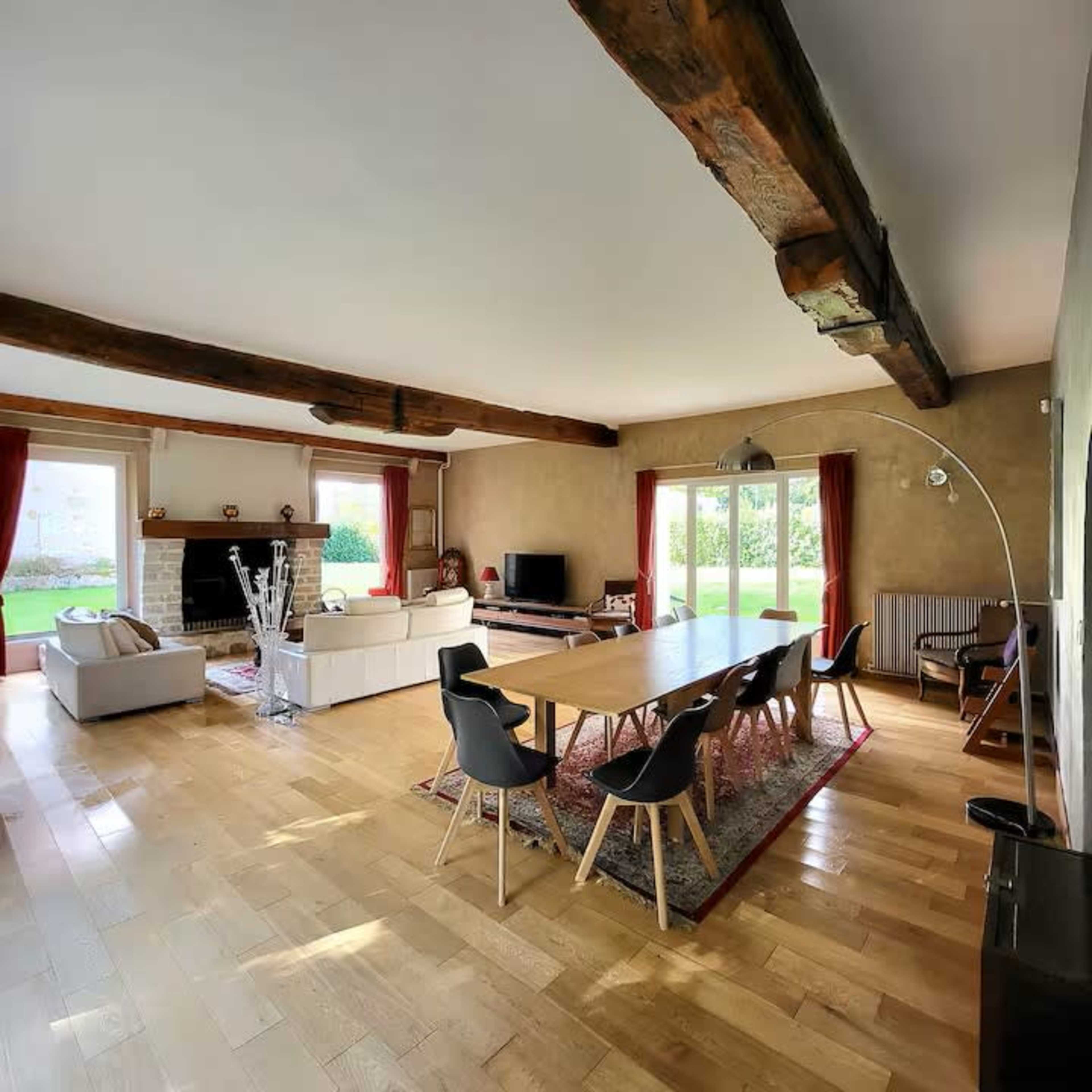 The image shows a spacious living room featuring a long dining table, modern chairs, a fireplace, and large glass doors leading to an outdoor area.