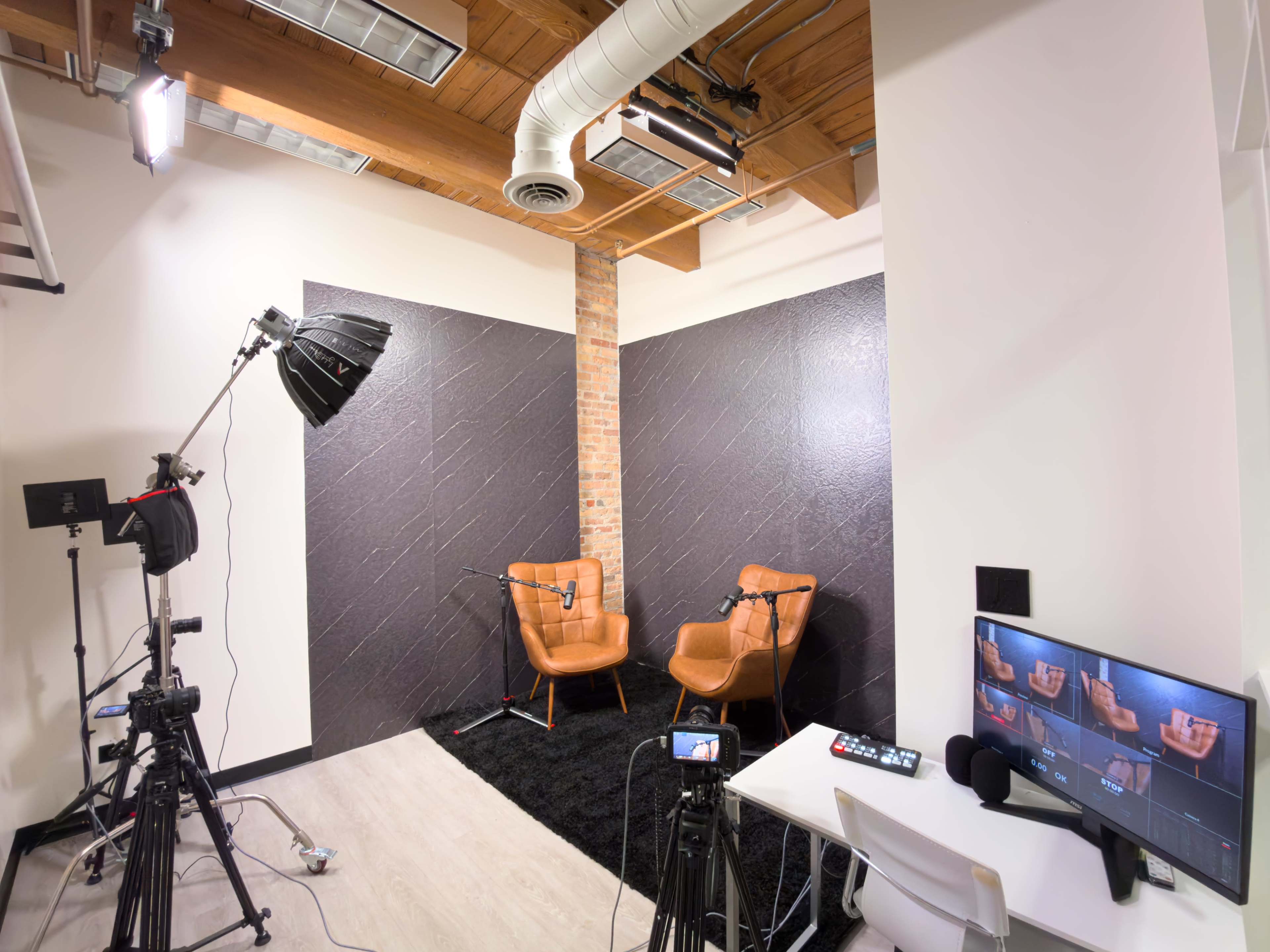 The image shows a video recording studio with two brown chairs against a textured black wall, equipped with lighting and camera equipment.