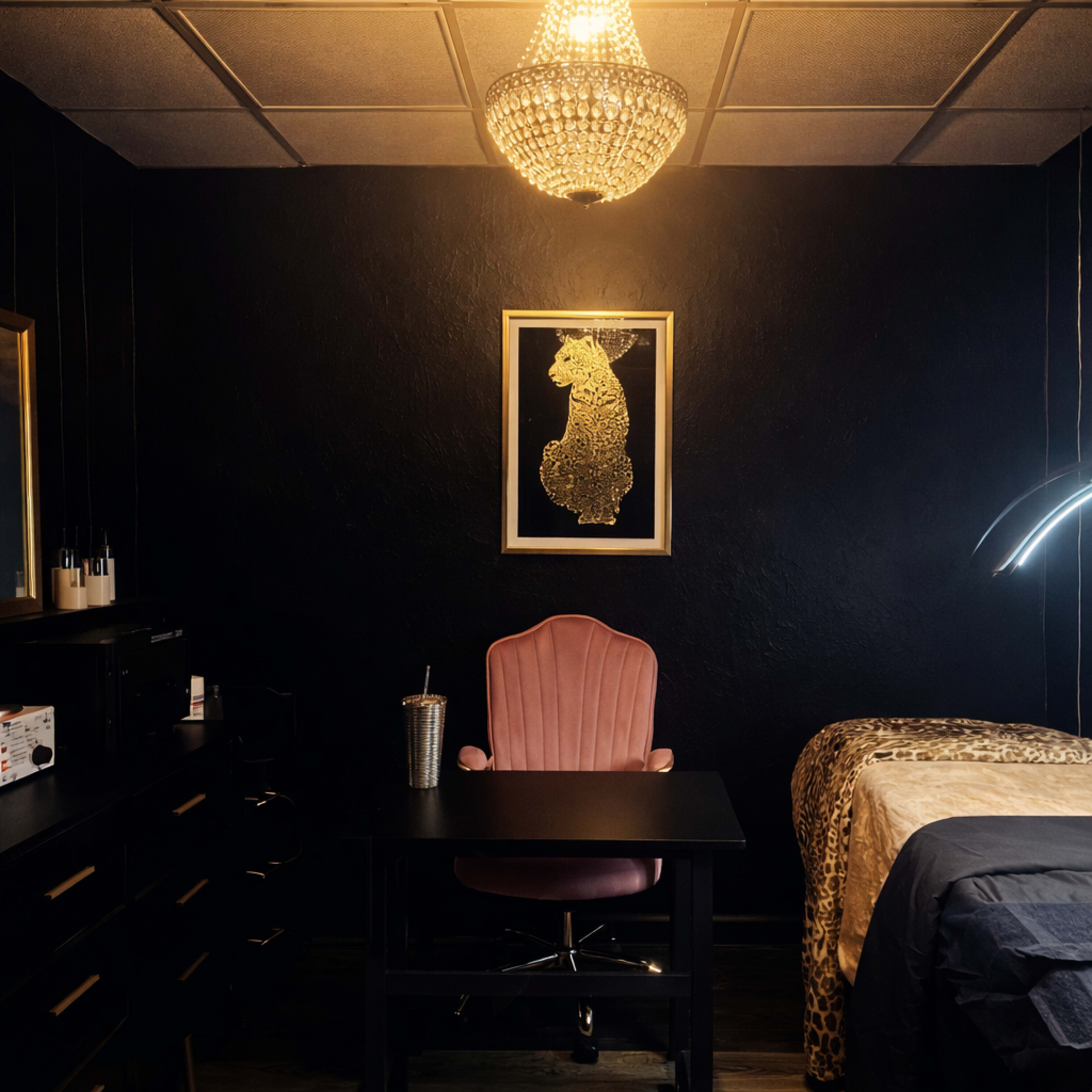 The image shows a dimly lit room with a black feature wall, a pink velvet chair at a desk, and a bed covered with a gray blanket, accented by a chandelier and a framed artwork of a lion.