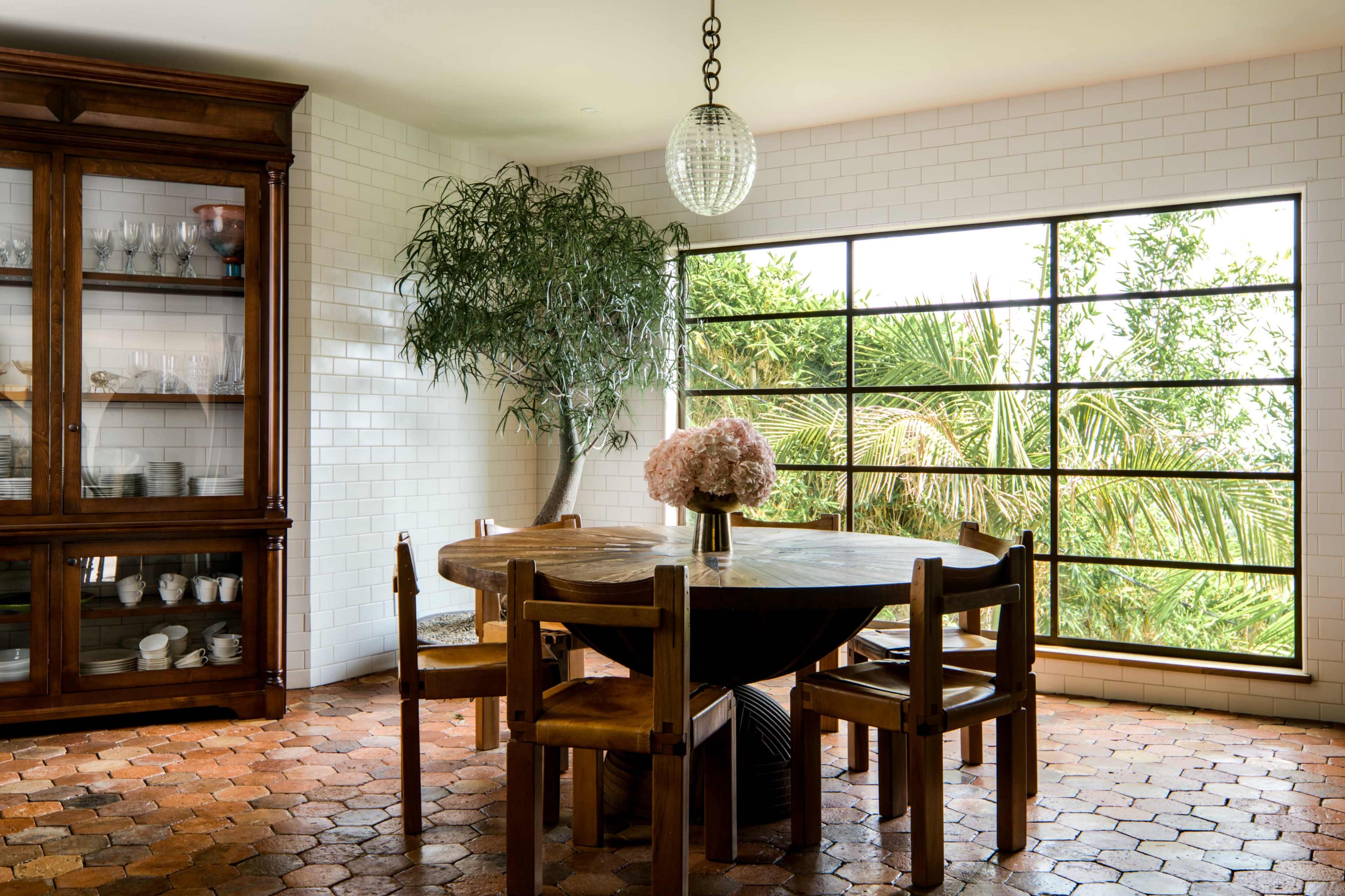 A dining area features a circular wooden table surrounded by chairs, with a large glass window overlooking greenery and a decorative potted tree.