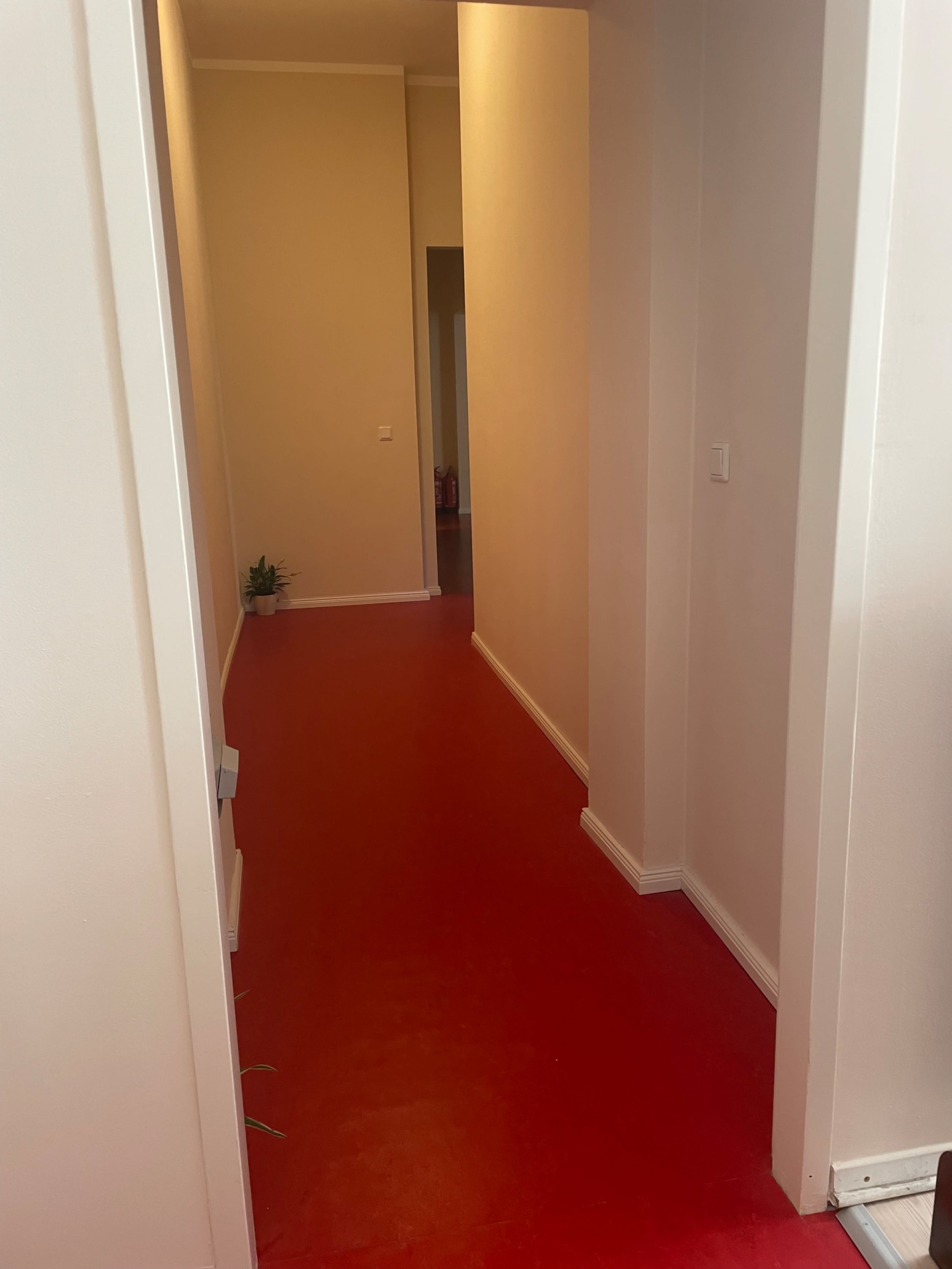 The image shows a hallway with red flooring and beige walls, leading to additional rooms at the far end.