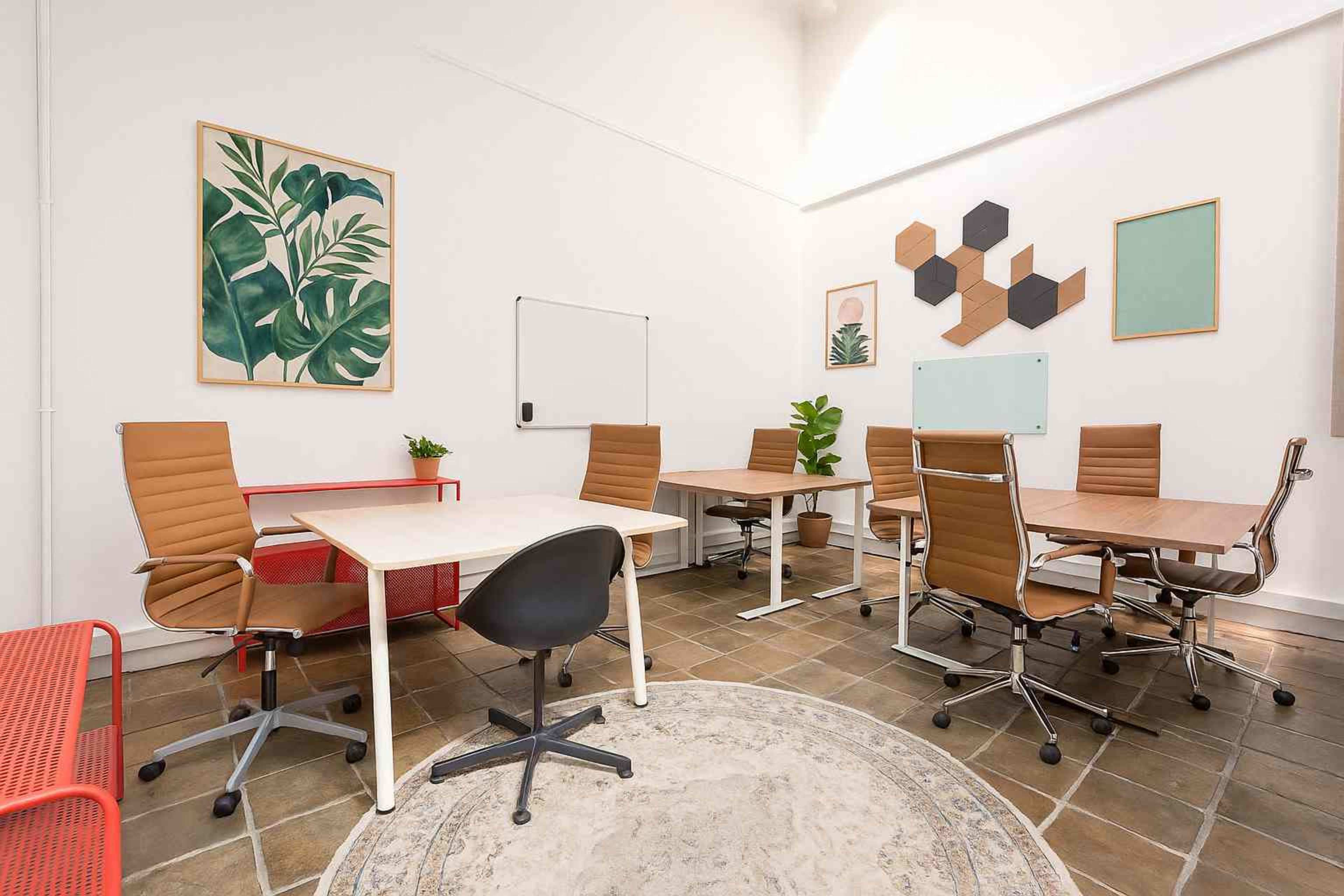 The image shows a modern, well-lit office space featuring several desks with ergonomic chairs, plants, and wall art.