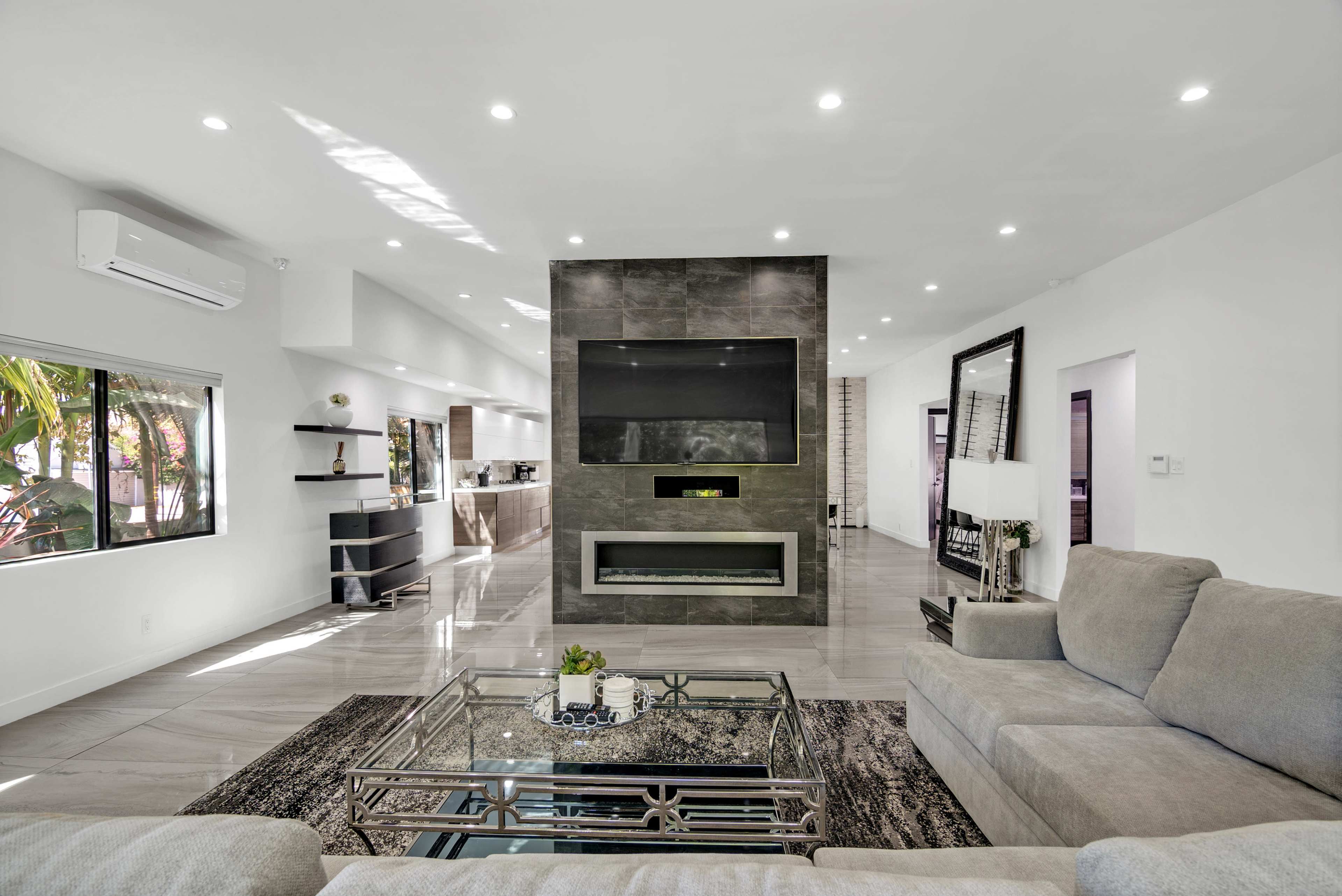 A modern living room features a large gray sectional sofa, a metallic coffee table, and a wall-mounted television above a sleek fireplace.