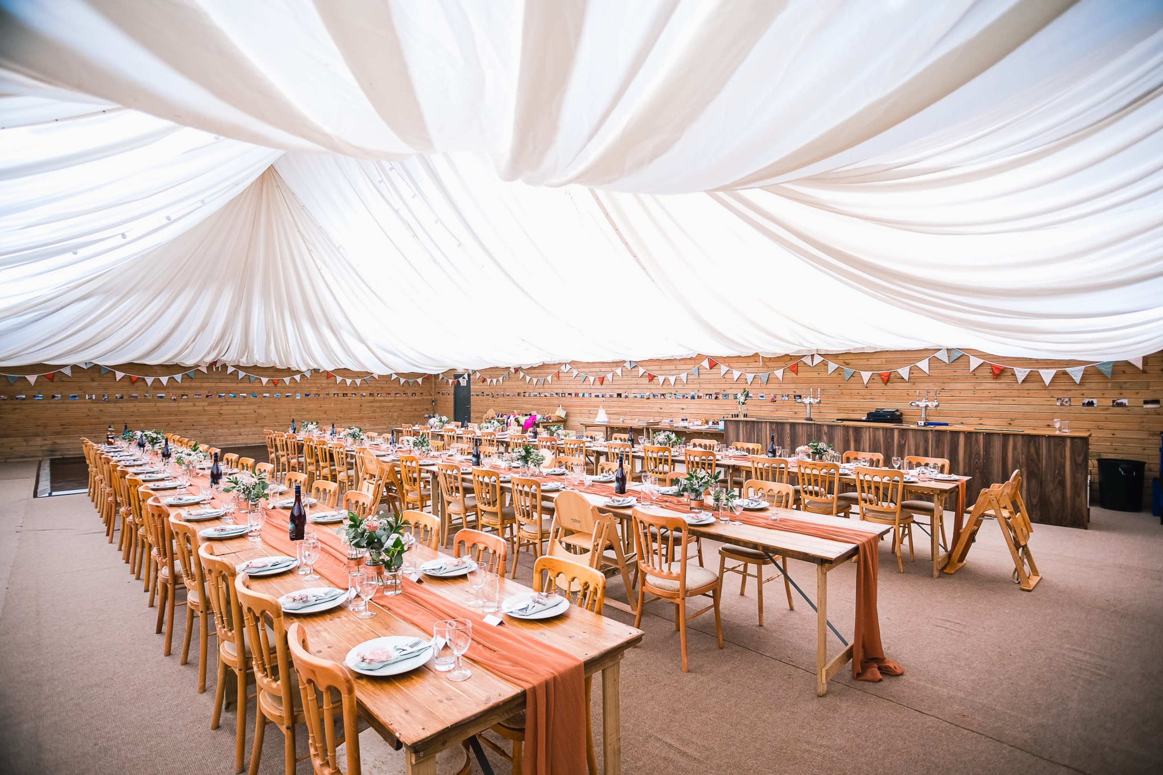 A large dining space is arranged with wooden tables and chairs under a draped white ceiling, decorated with floral centerpieces and bunting.