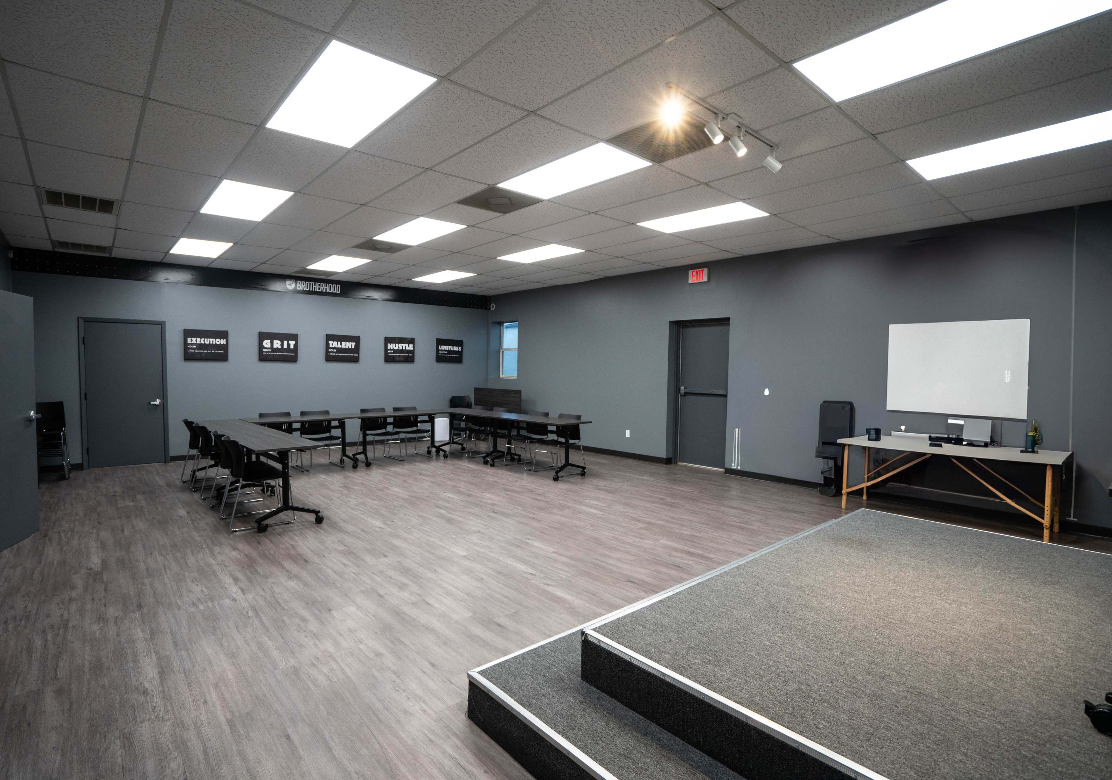 The image shows a spacious, well-lit meeting room featuring rows of tables and chairs arranged around a small stage area.