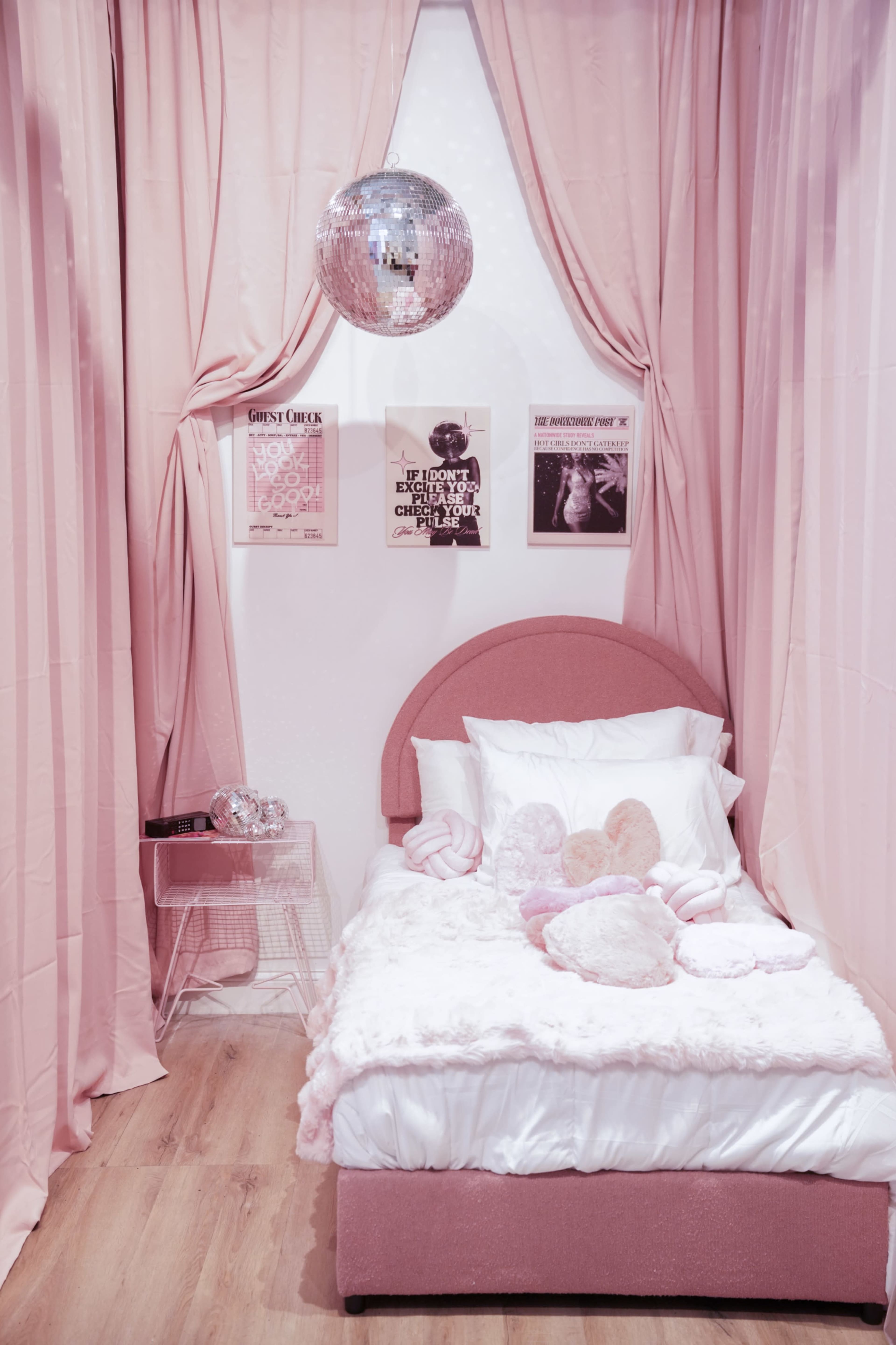 The image shows a cozy bedroom featuring a pink bed adorned with soft pillows and plush toys, surrounded by pink curtains and decorated with framed posters on the wall.