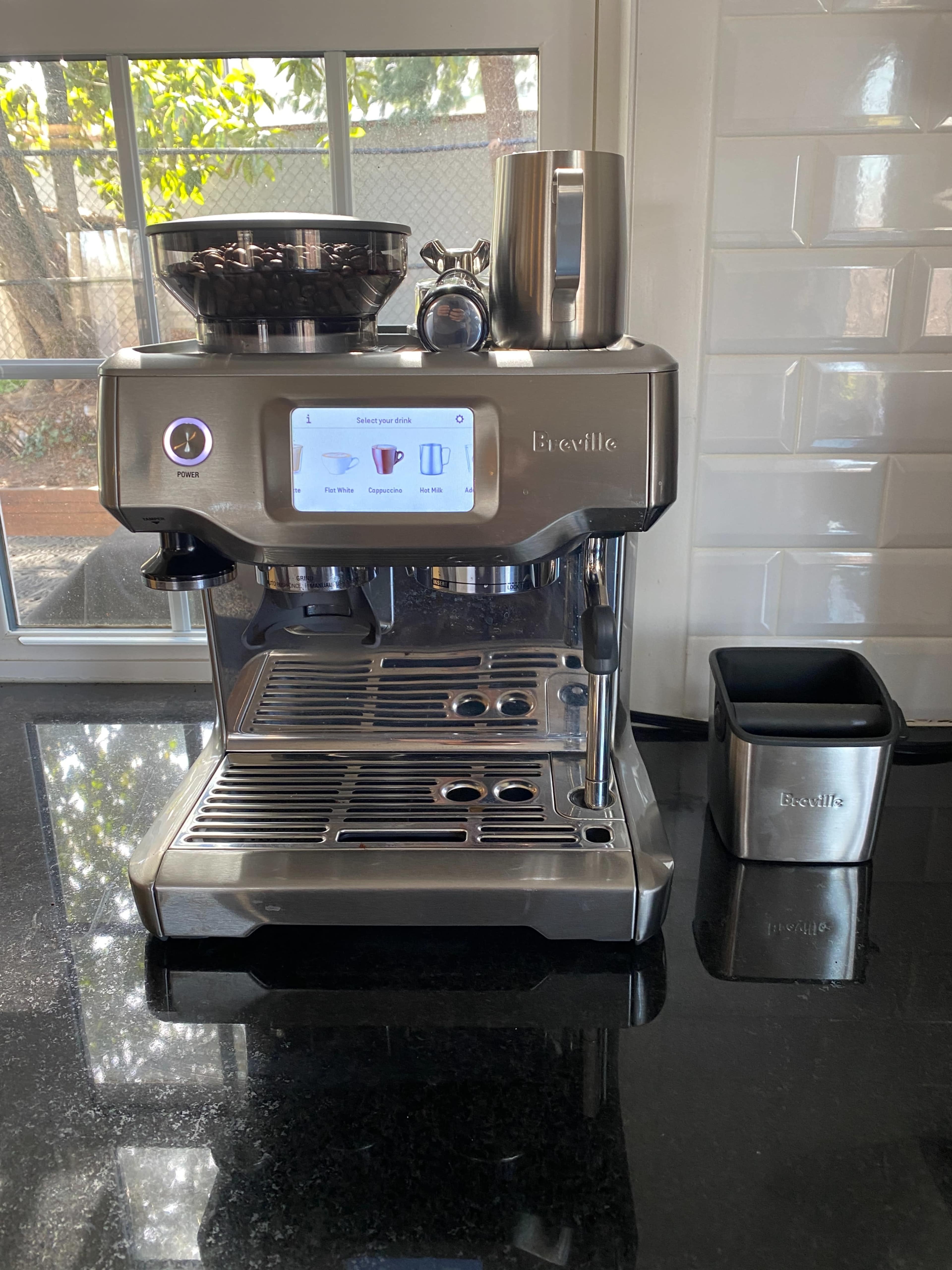 A stainless steel Breville espresso machine is positioned on a black countertop, with a clear display screen and various accessories nearby.