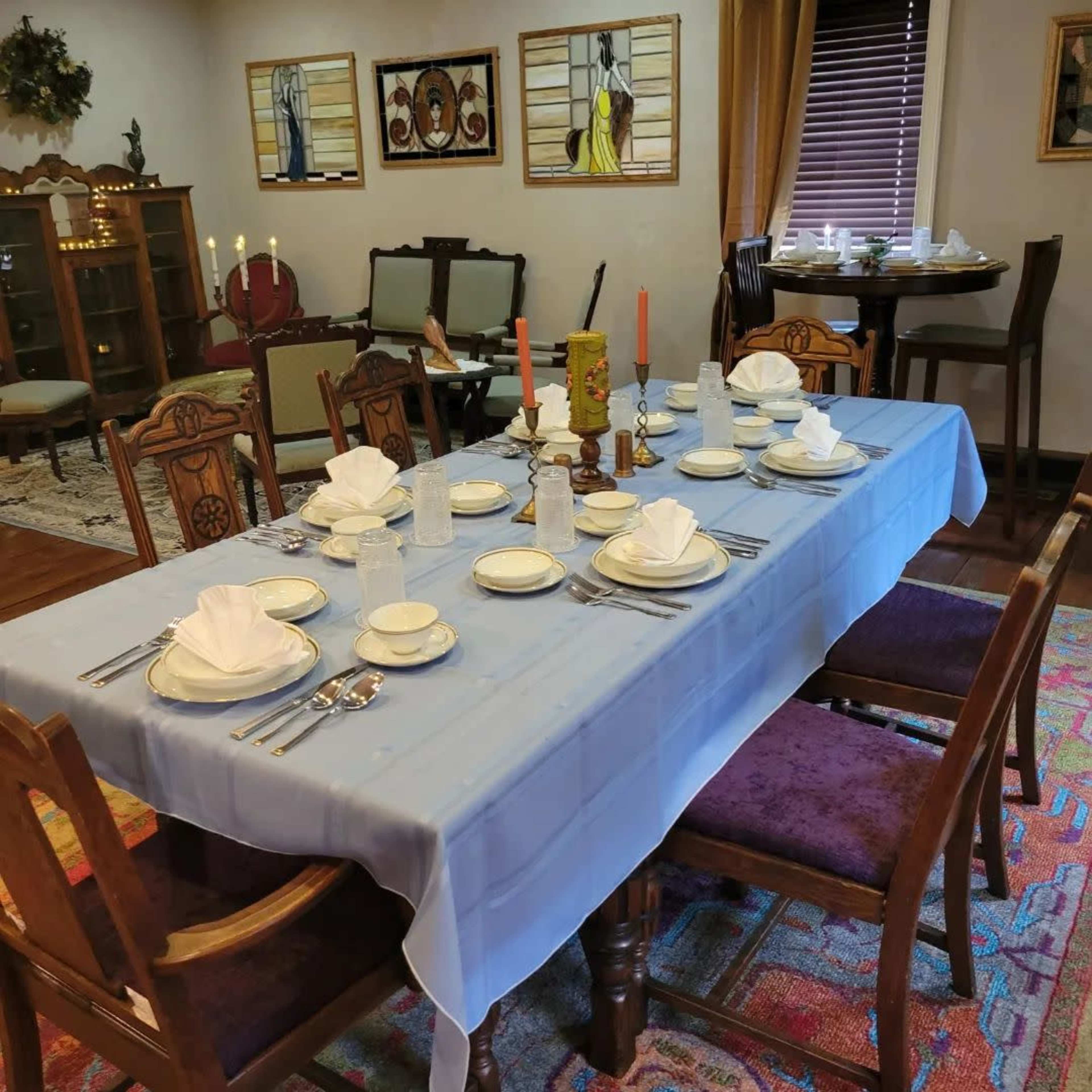 A dining room table is set for a meal with white plates, folded napkins, glasses, and silverware, surrounded by wooden chairs against a backdrop of decorated walls and furniture.