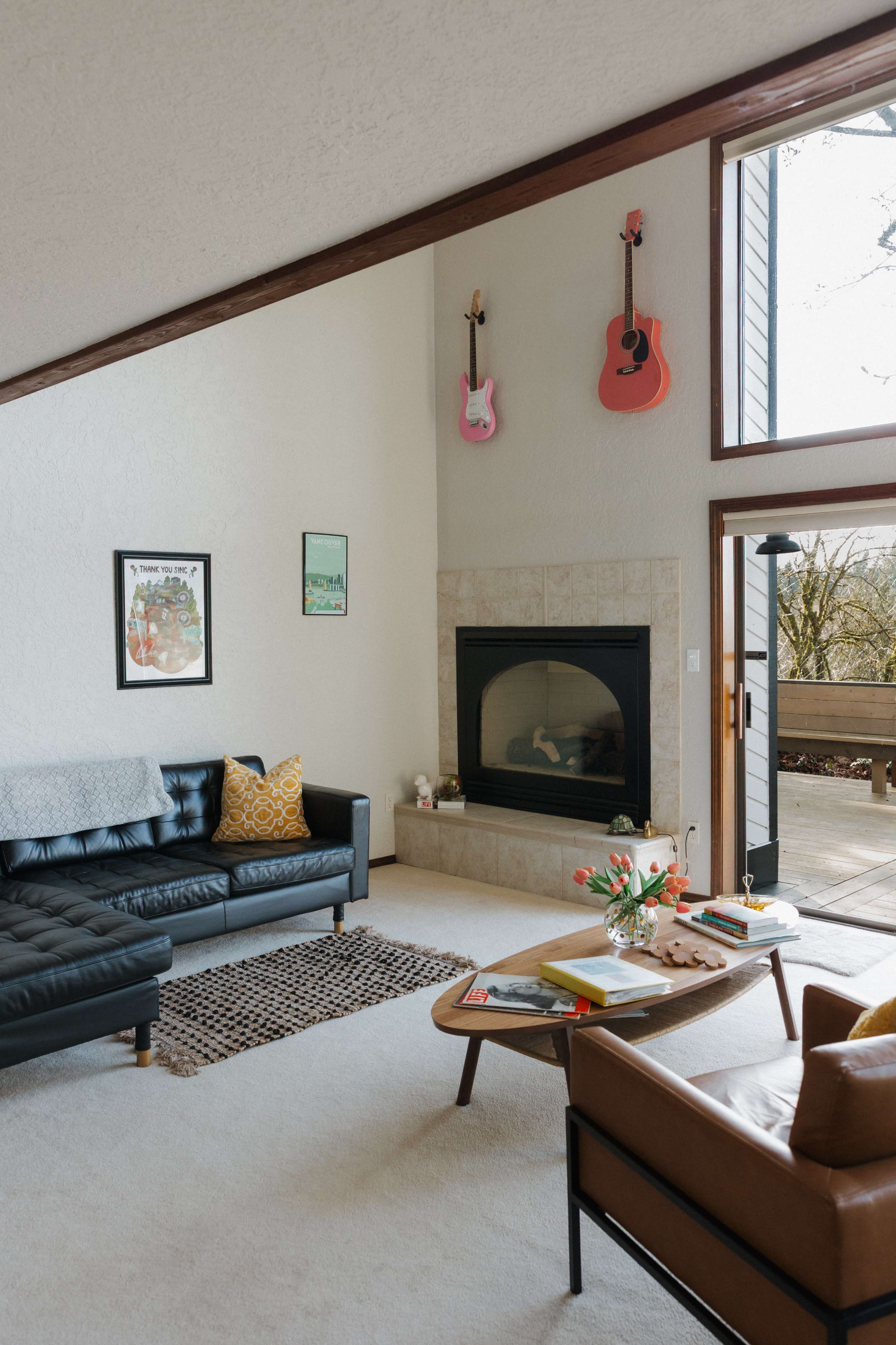 The living room features a black leather sofa, a coffee table, a fireplace, and light streaming in through large windows.