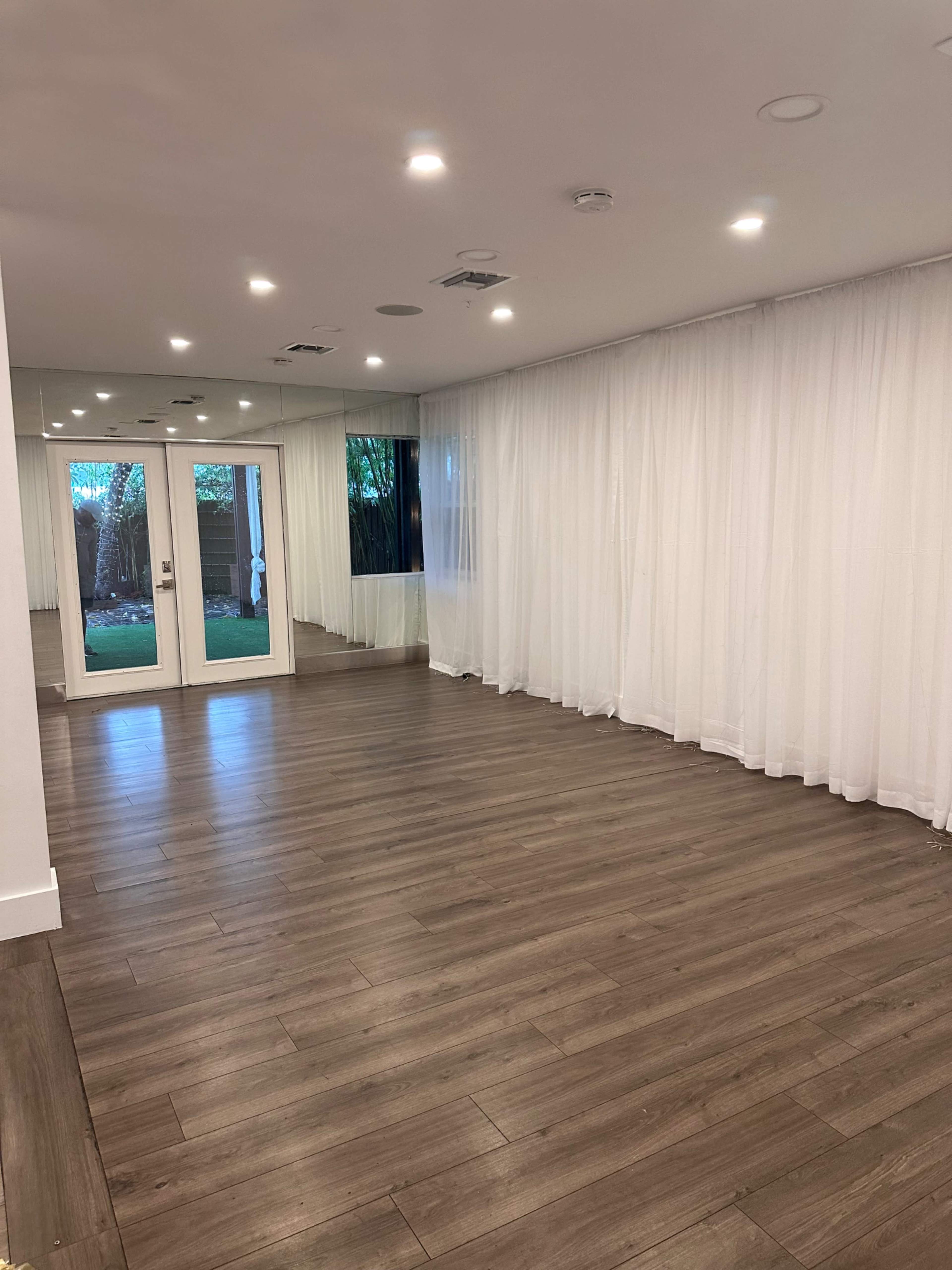 An empty room with wooden flooring, large windows with white sheer curtains, and double doors leading outside.