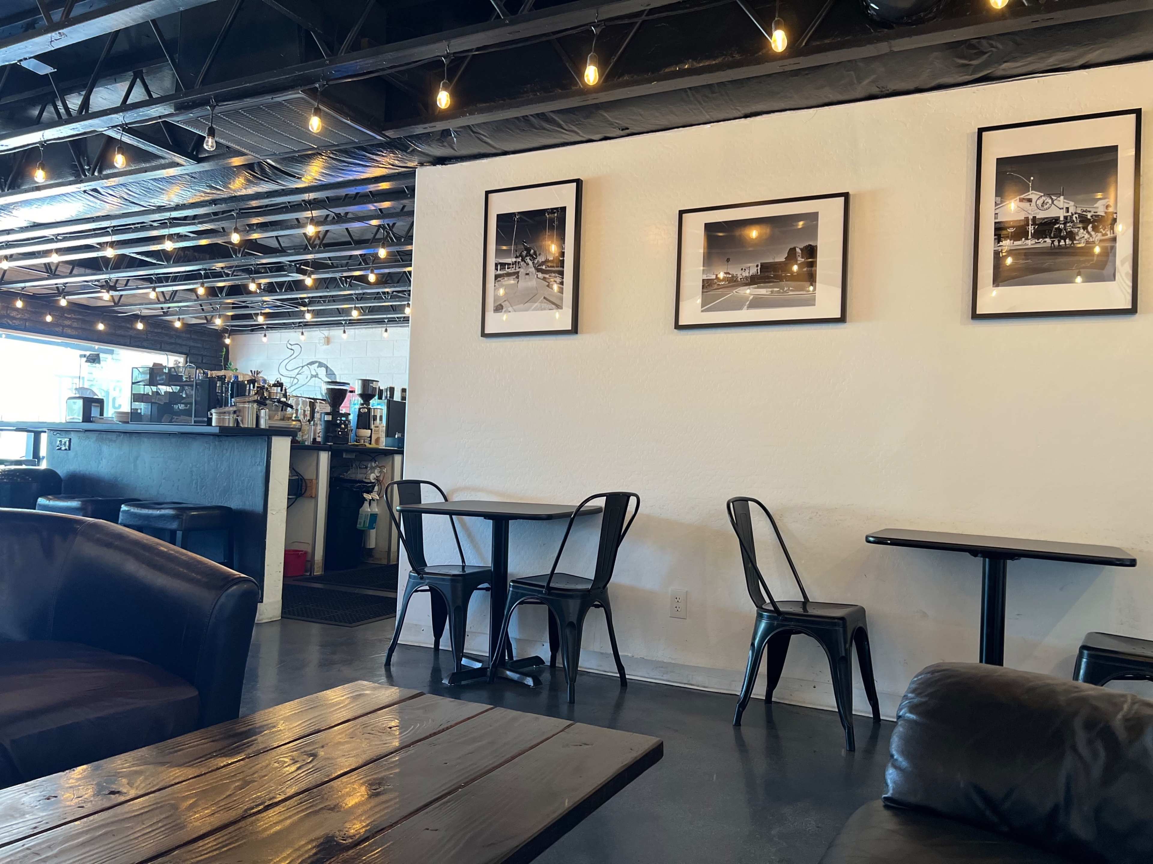 A cozy cafe interior with a few tables and chairs, framed photographs on the wall, and string lights overhead.