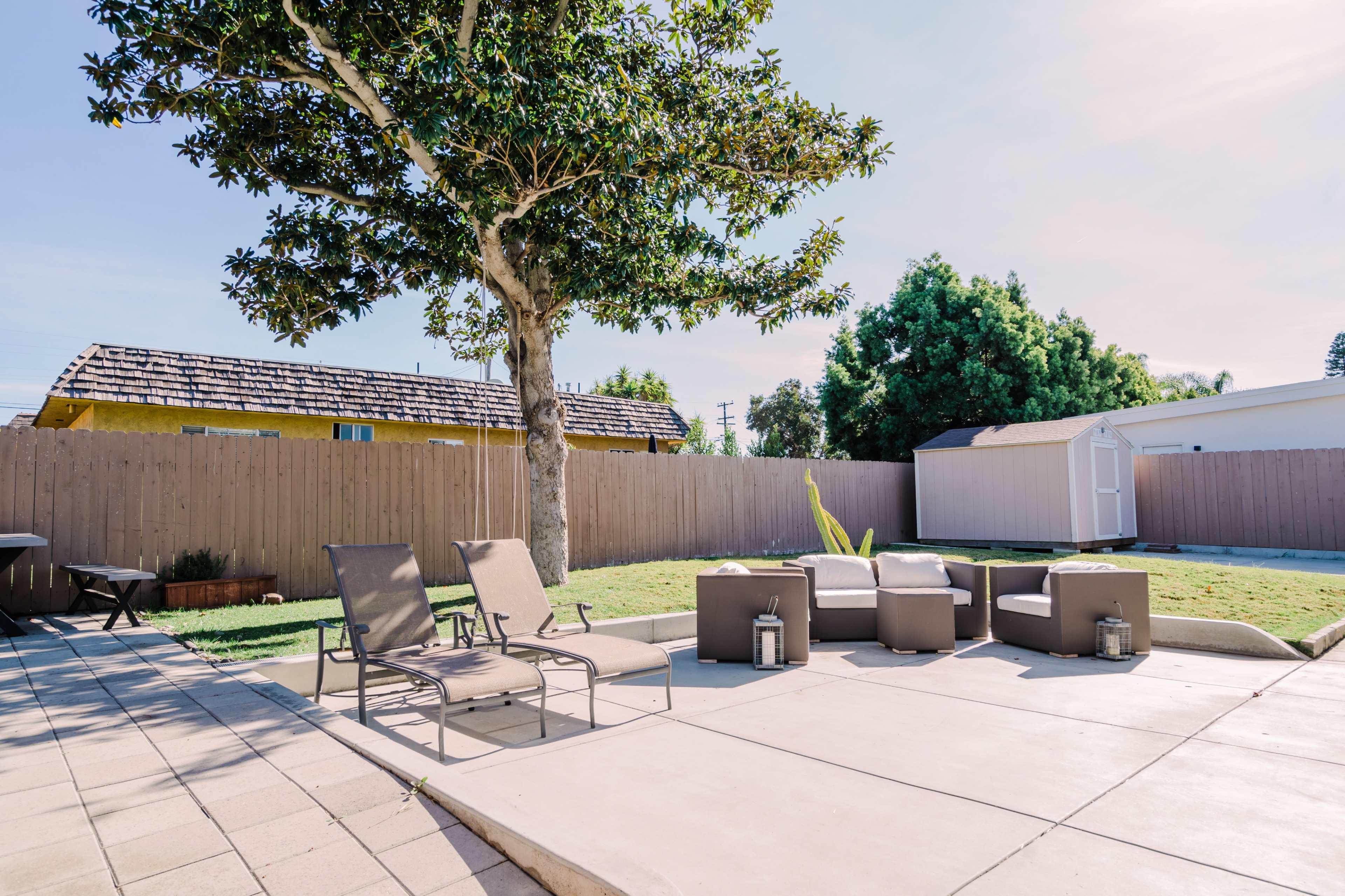 The image shows a backyard setting with patio furniture arranged on a concrete surface, surrounded by a wooden fence and a tree.