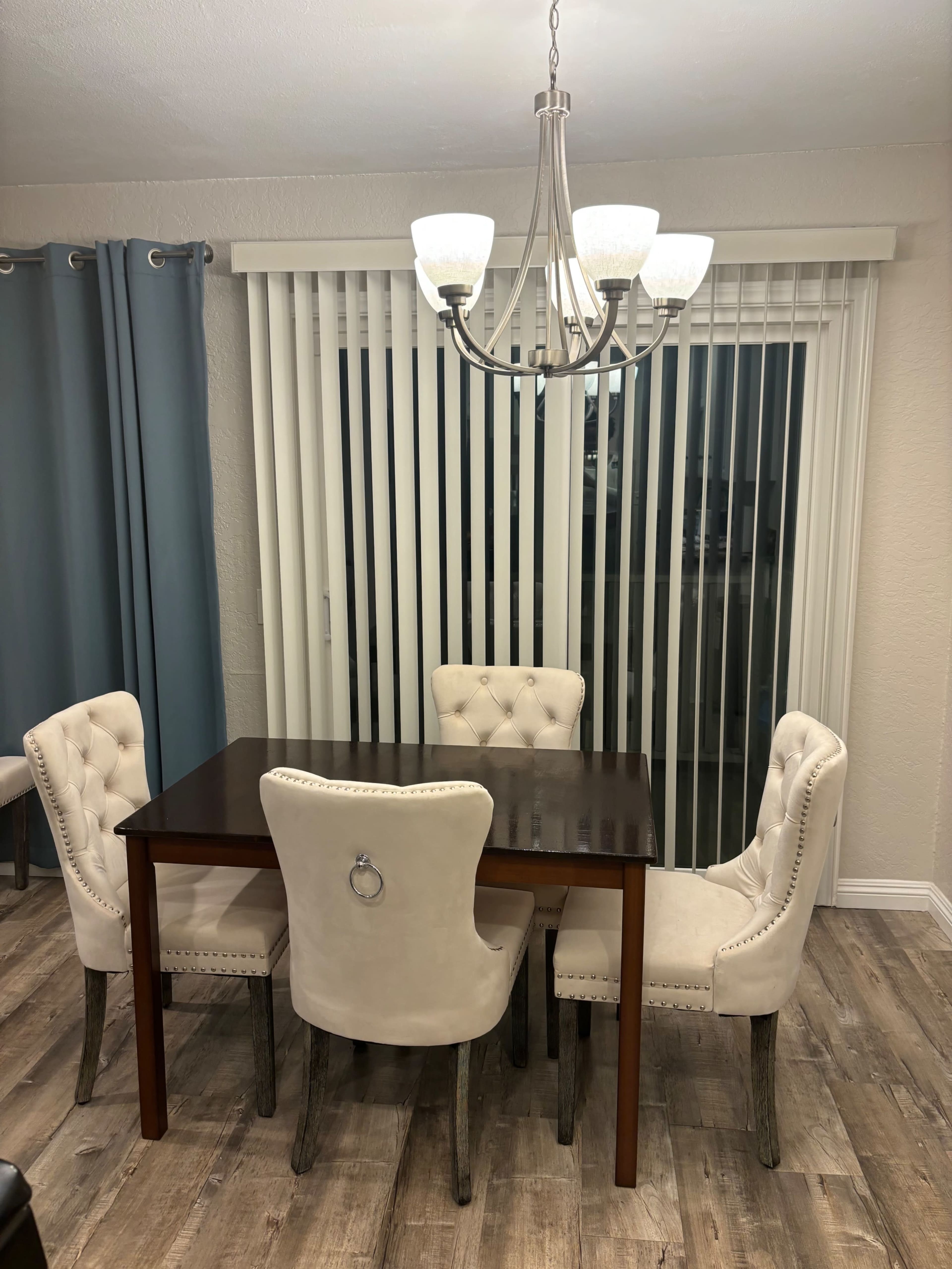 A wooden dining table is surrounded by four upholstered chairs beneath a chandelier, with vertical blinds covering a large window in the background.