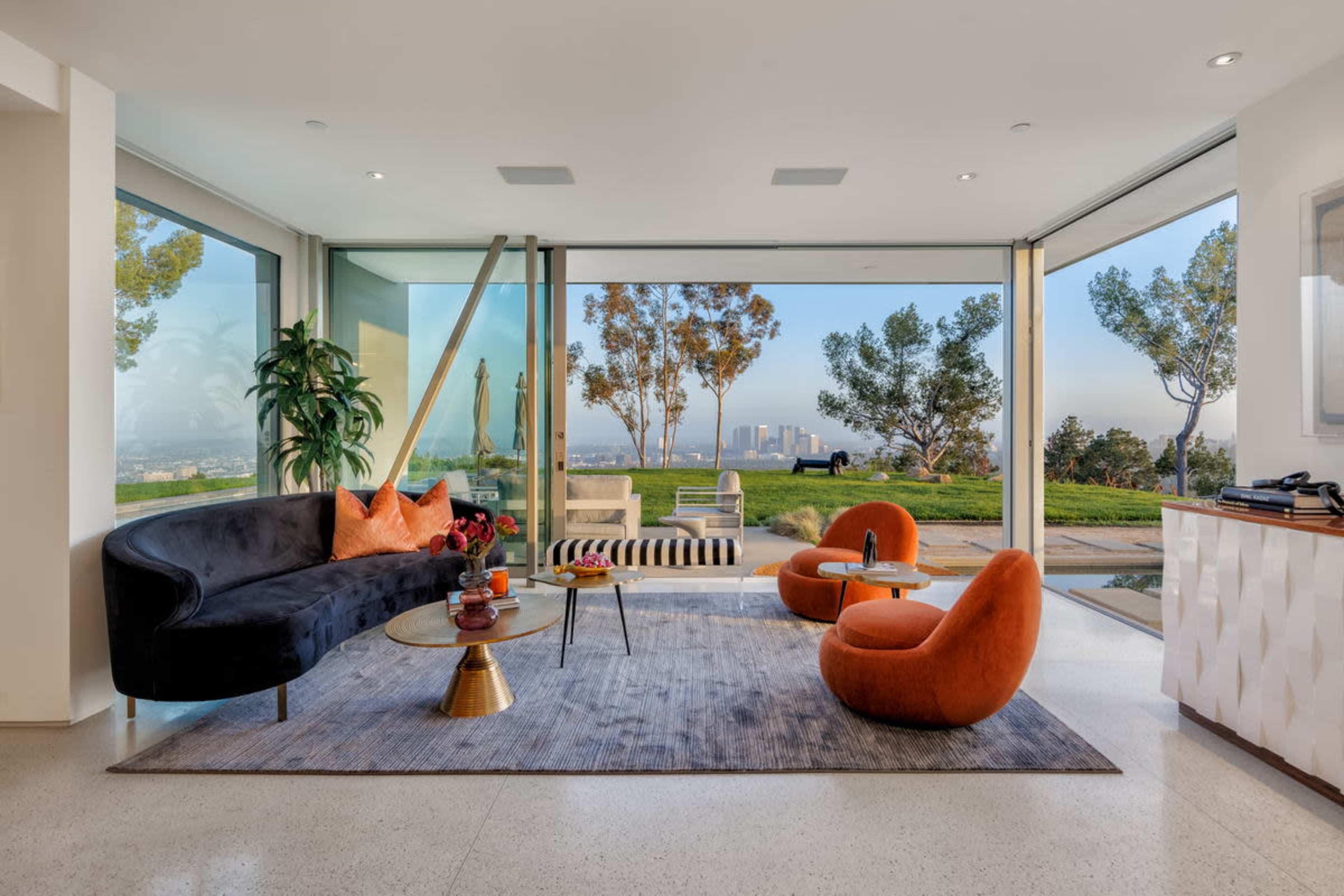 A modern living room features a black sofa, orange chairs, a glass coffee table, and large windows showcasing a city view and surrounding greenery.