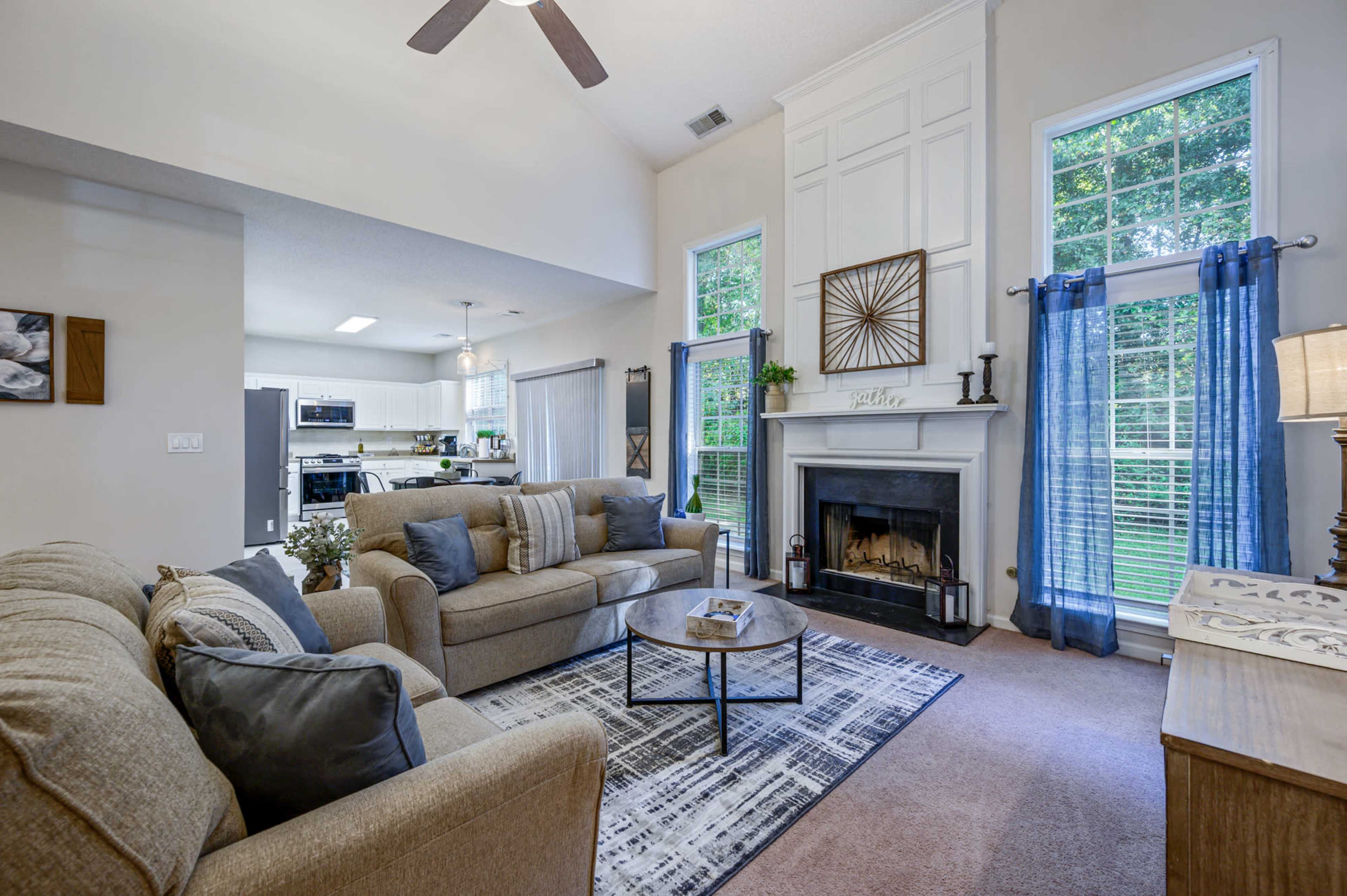 A living room with a beige sofa and a matching armchair, a round coffee table in the center, and a fireplace against a wall with large windows.