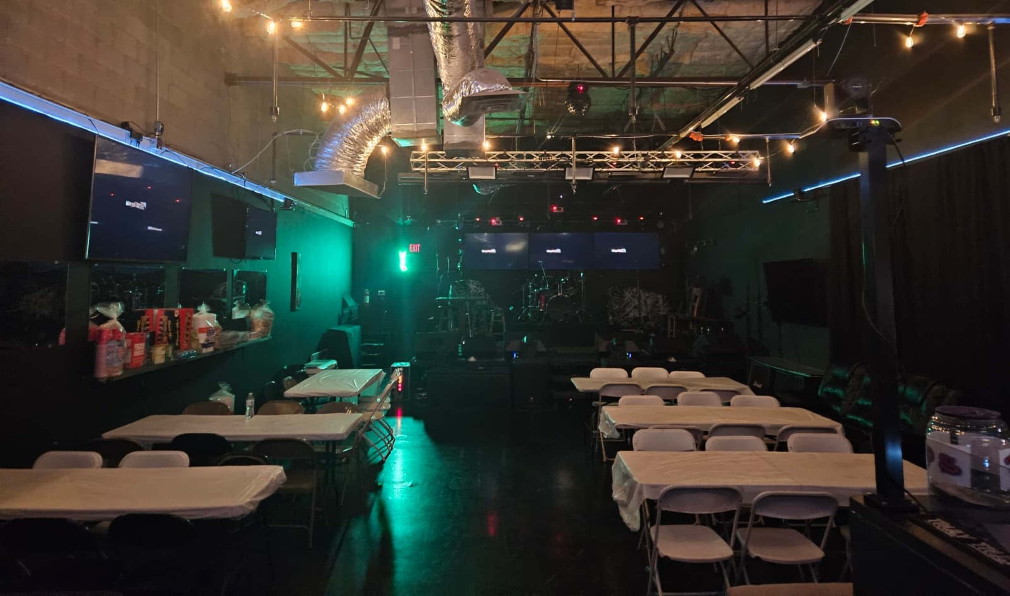 The image shows a dimly lit venue with a stage at the front, surrounded by tables and chairs, and illuminated by green and blue lights.
