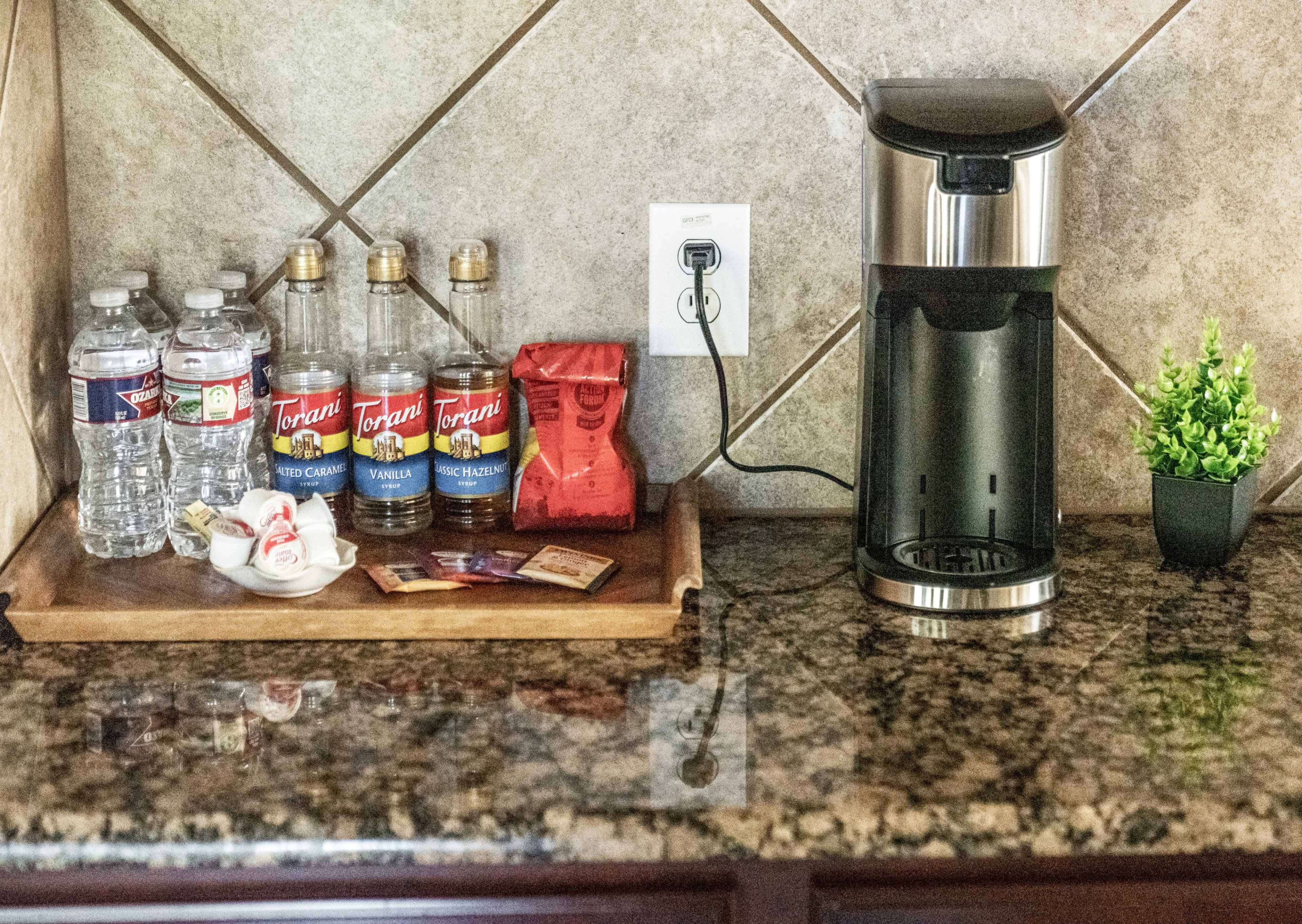 A kitchen counter with bottles of flavored syrup, water bottles, coffee pods, sugar packets, and a coffee maker on a granite surface.