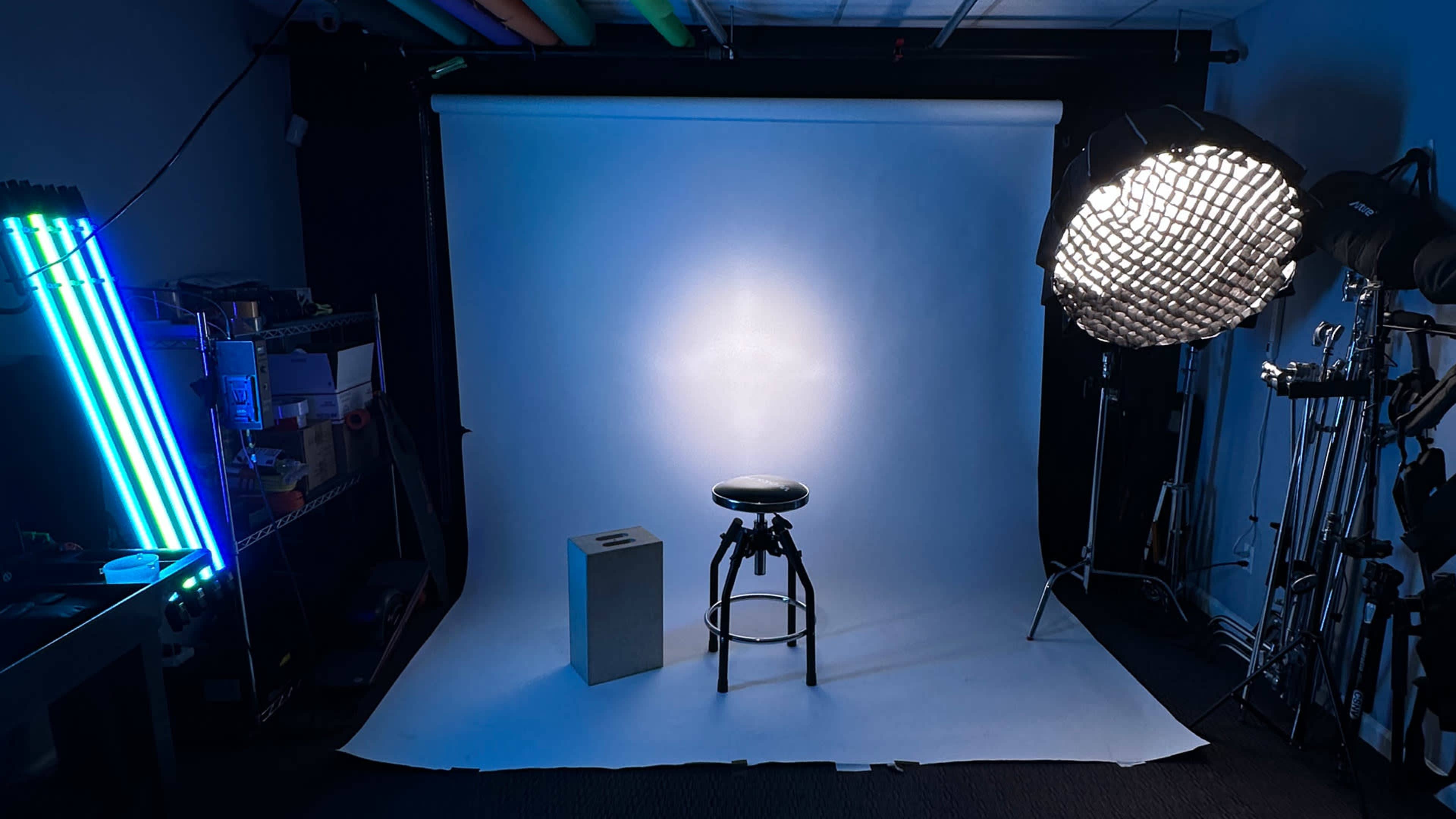 A photography studio setup featuring a black stool and a gray cube in front of a white backdrop, illuminated by a large softbox and accented by colorful LED lights on the side.
