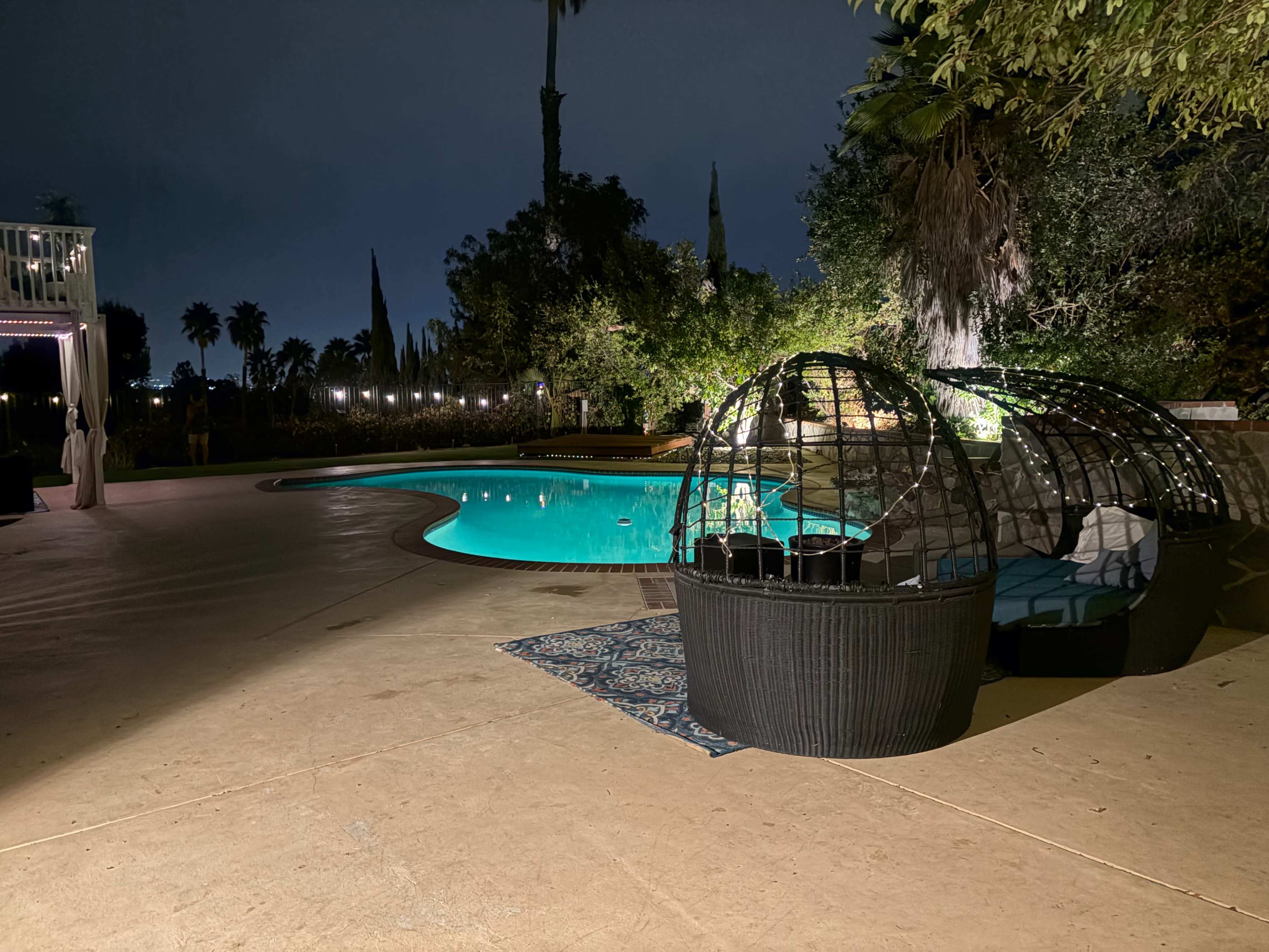 The scene features a well-lit outdoor pool area at night, with a pair of dome-shaped seating structures beside the pool, surrounded by greenery and palm trees.