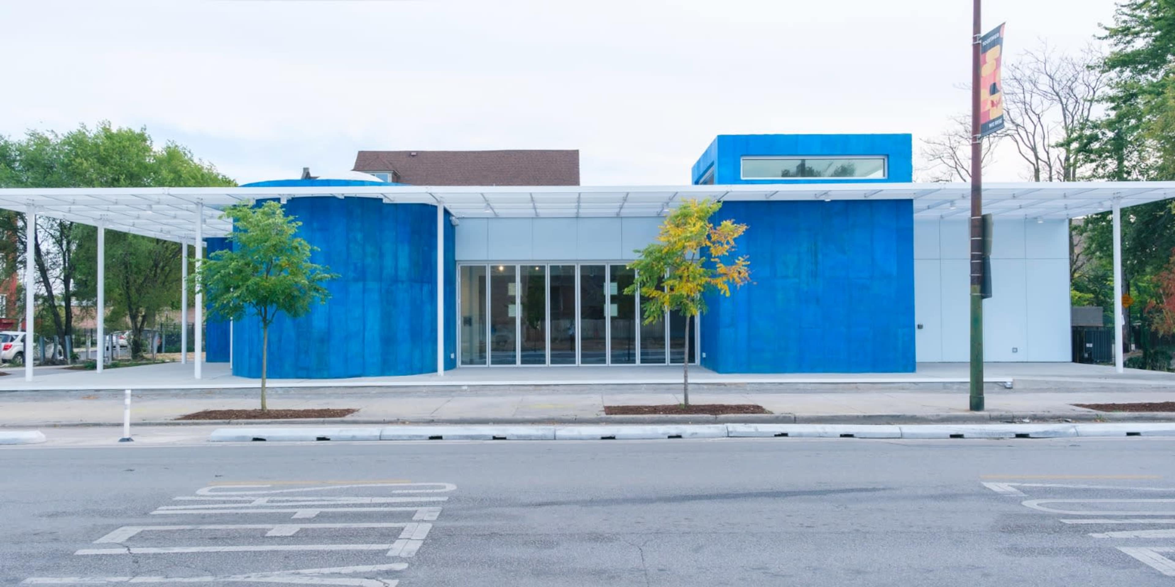 The image shows a modern, blue-clad building with a covered entrance and trees on either side, situated along a street.