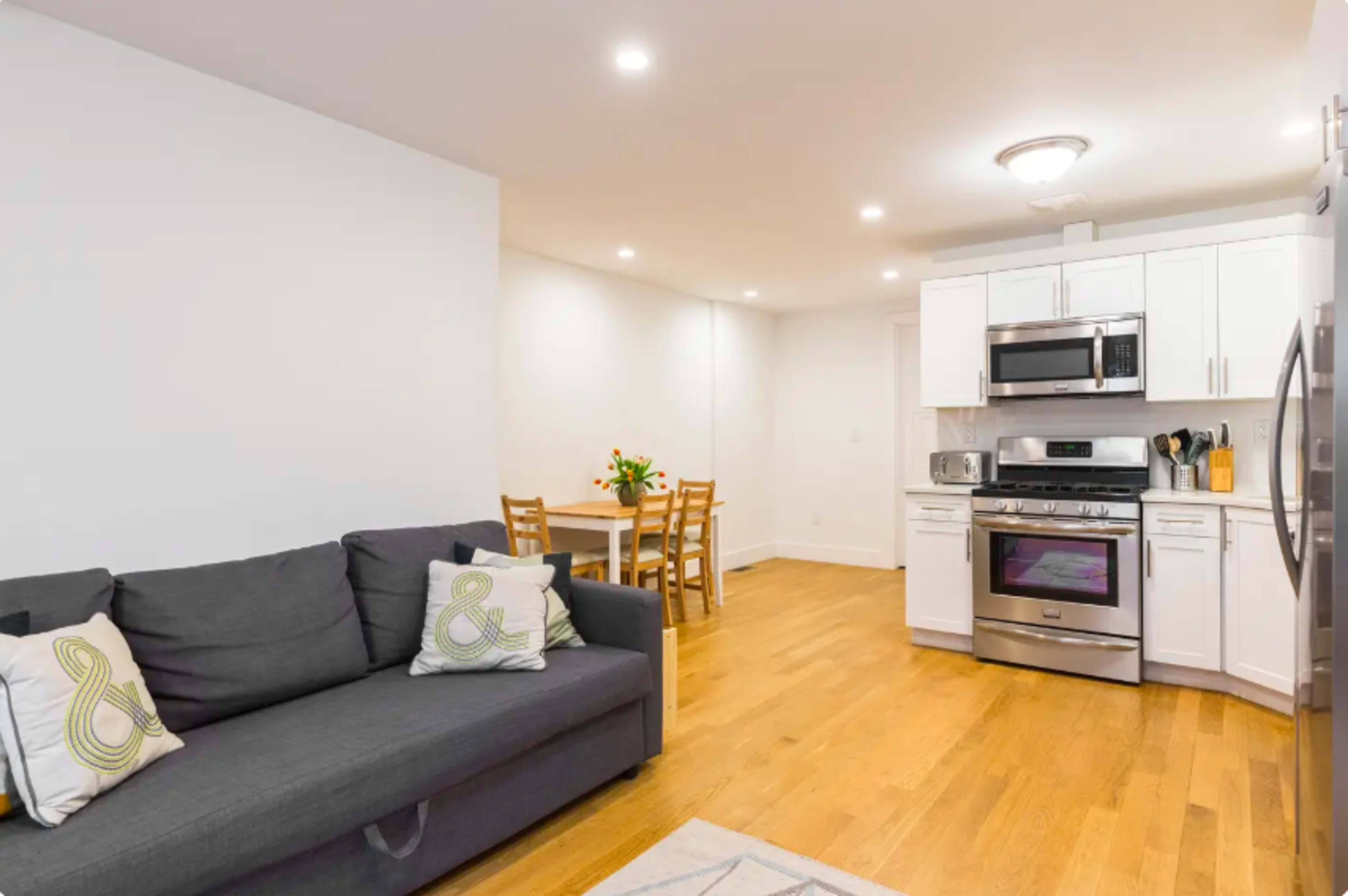 A modern kitchen and living area with a gray sofa, a dining table with chairs, and stainless steel kitchen appliances.