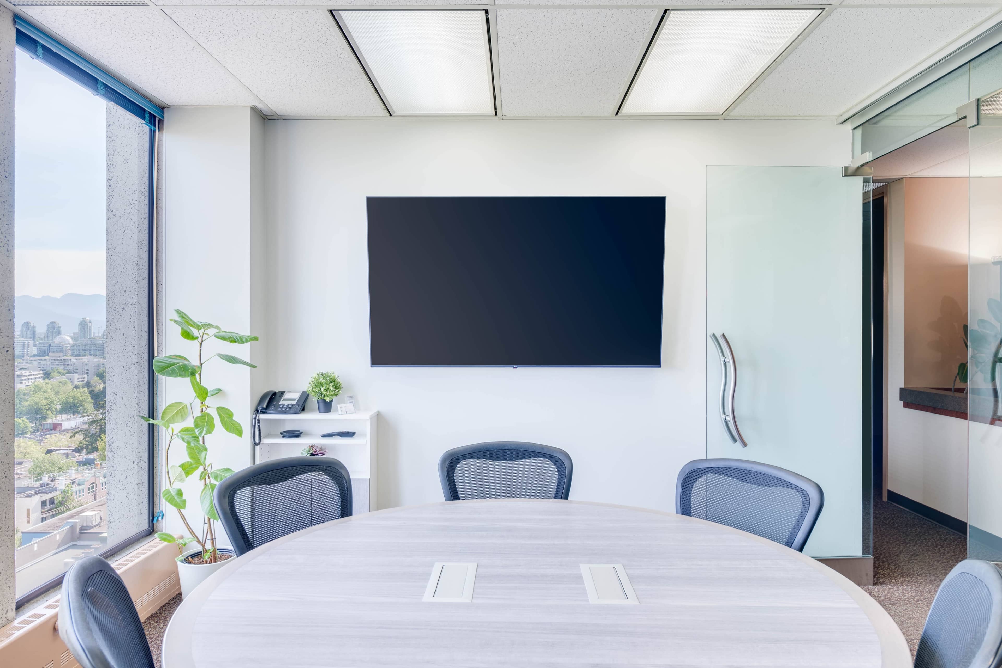 A modern conference room features a round table, several chairs, a wall-mounted television, and a large window with a city view.